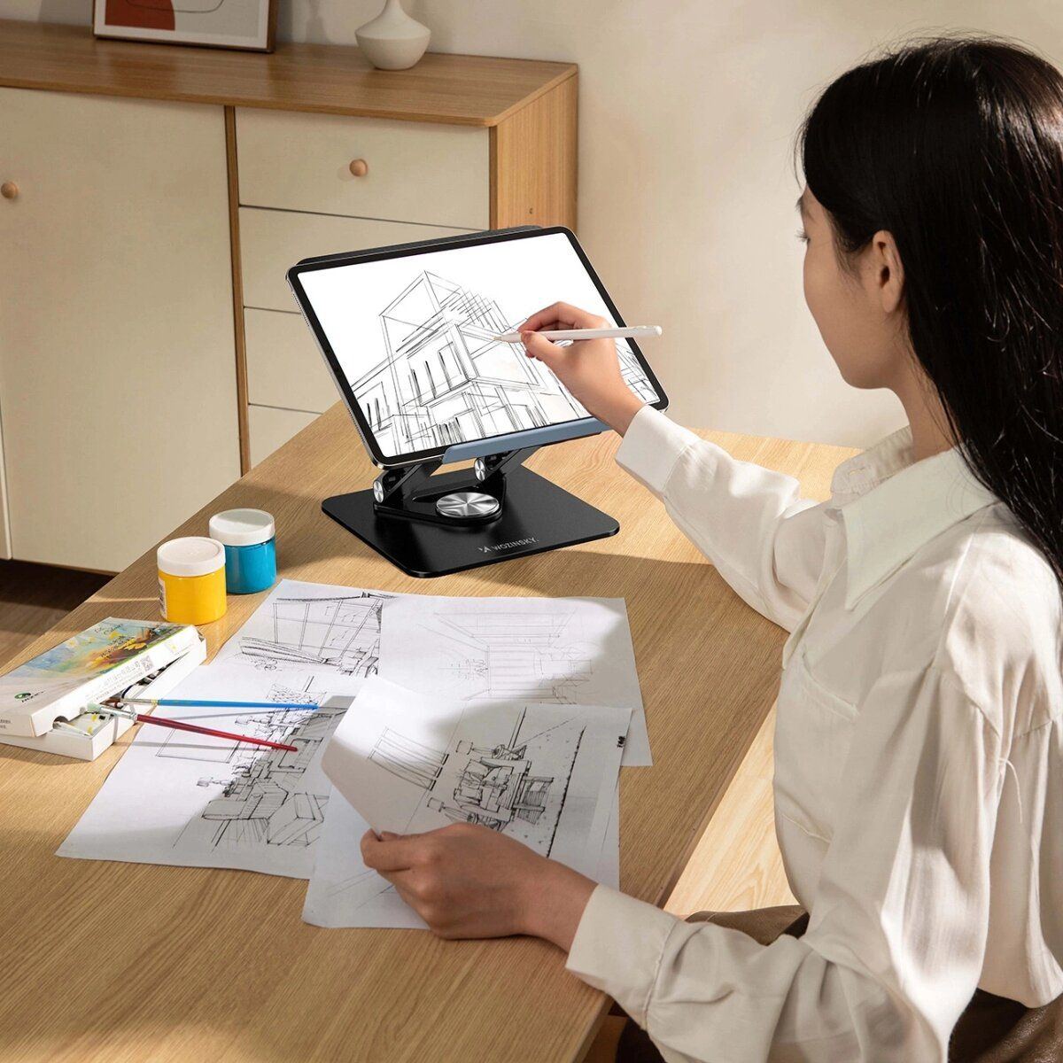 Kobieta rysująca na tablecie. Tablet na stojaku na biurku. Widoczna farba i rysunki.