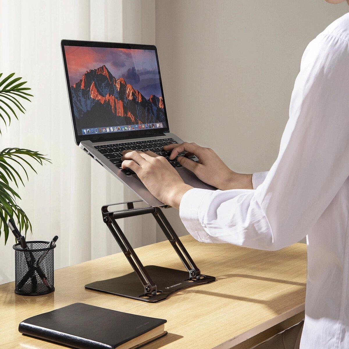 Eine Person, die einen Laptop auf einem schwarzen, verstellbaren Ständer benutzt. Schreibtisch mit Notizbuch und Stift.