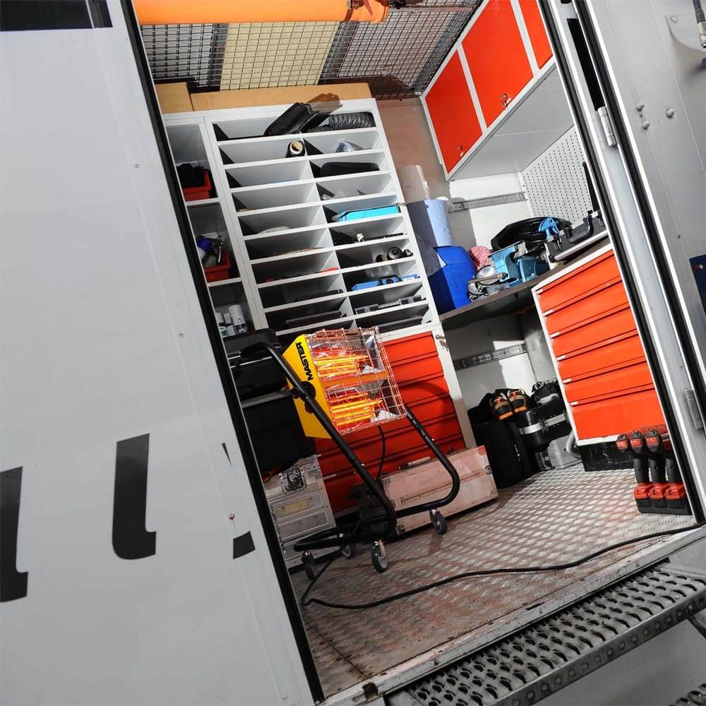 Binnen in een vrachtwagen: planken, laden en een kachel. Rode laden, zilveren vloer en witte muren.