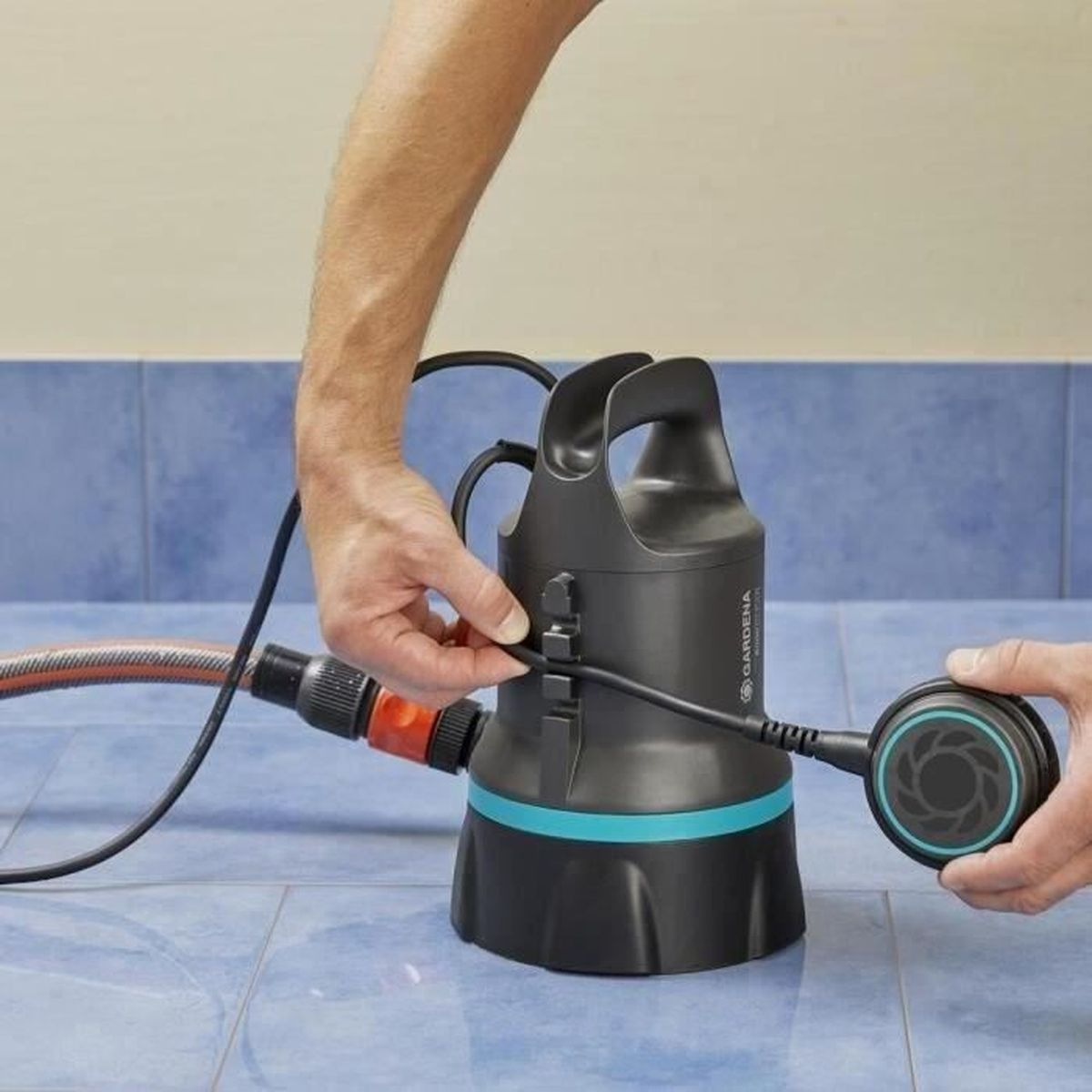 Le mani collegano un tubo a una pompa nera. Pavimento piastrellato blu. La pompa ha una maniglia e un cavo nero.
