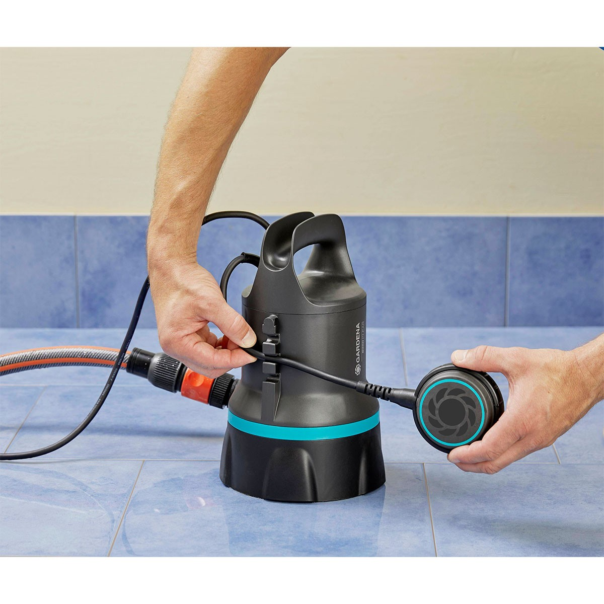 Una persona collega un tubo a una pompa dell'acqua nera e verde acqua su un pavimento di piastrelle blu.