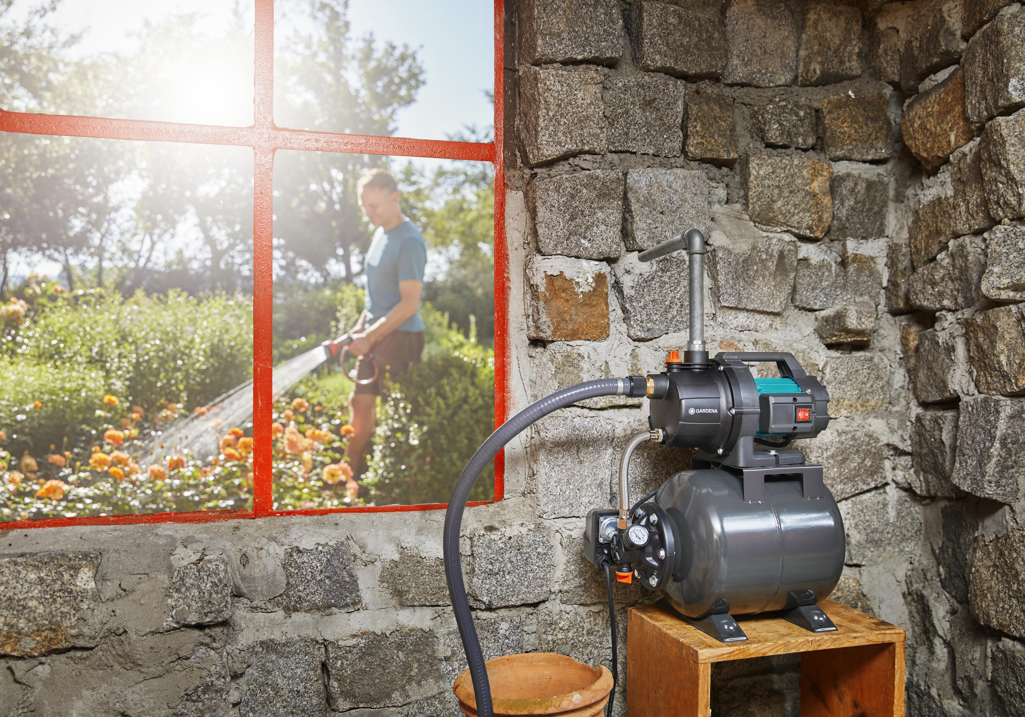 Eine Person bewässert einen Garten durch ein Fenster. Ein Pumpensystem befindet sich in einem Steingebäude.