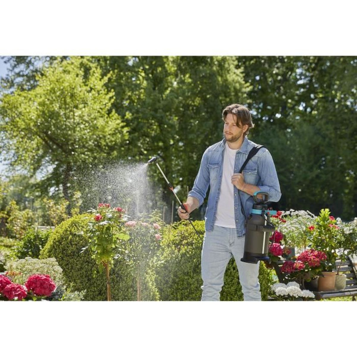 Mann besprüht Pflanzen im Garten. Er benutzt ein Sprühgerät. Grüne Bäume und rote Blumen im Hintergrund.