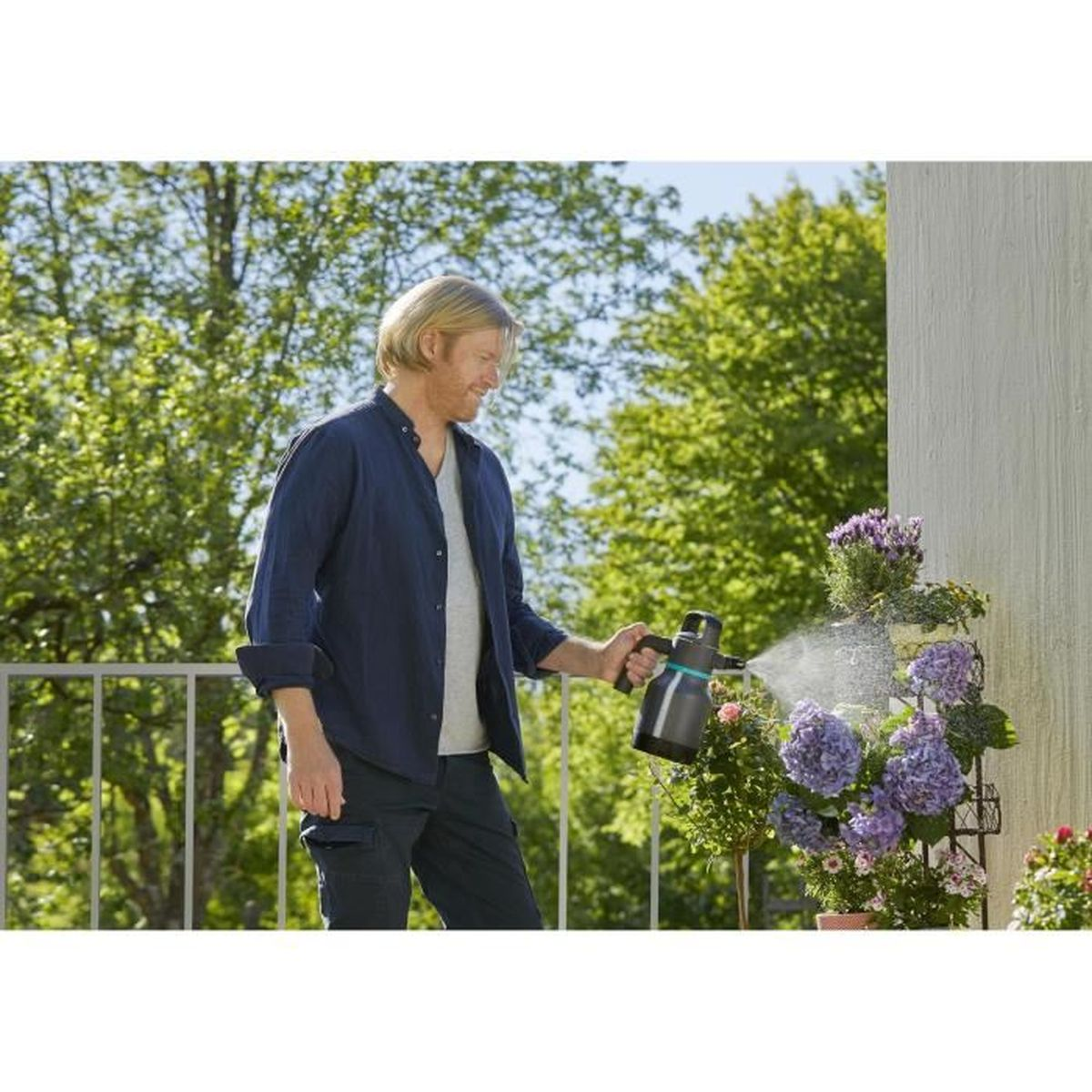 Mann sprüht Wasser auf lila Blumen auf einem Balkon. Grüne Bäume und Himmel im Hintergrund.