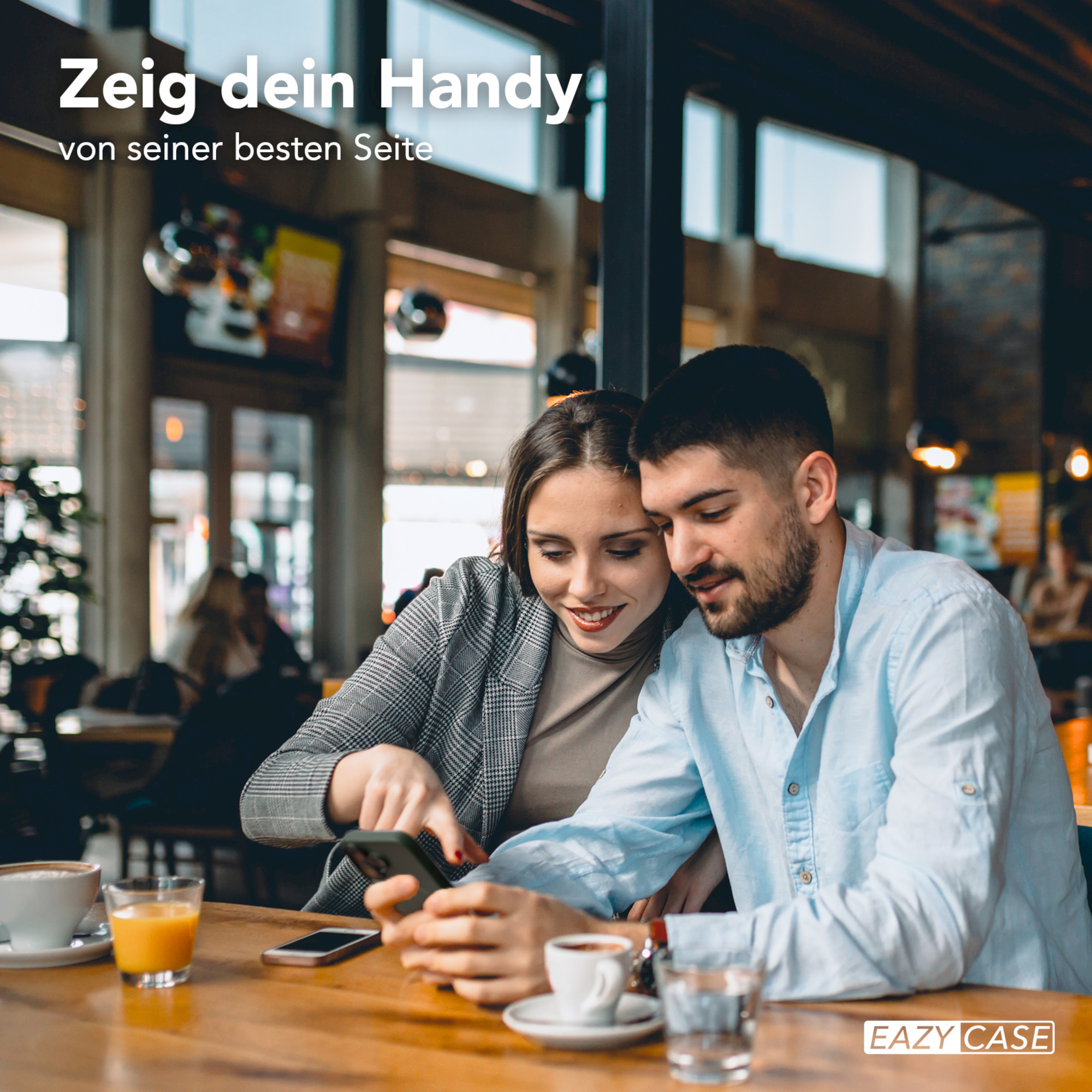 Ein Paar schaut in einem Café auf ein Telefon. Der Mann trägt ein blaues Hemd. Sie sitzen an einem Holztisch.