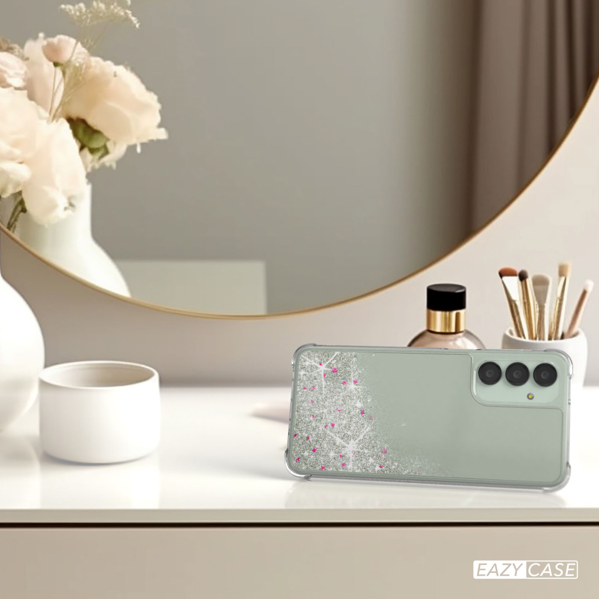 Eine Handyhülle auf einem Tisch mit Spiegel und Blumen im Hintergrund. Die Hülle hat Glitzer und rosa Akzente.