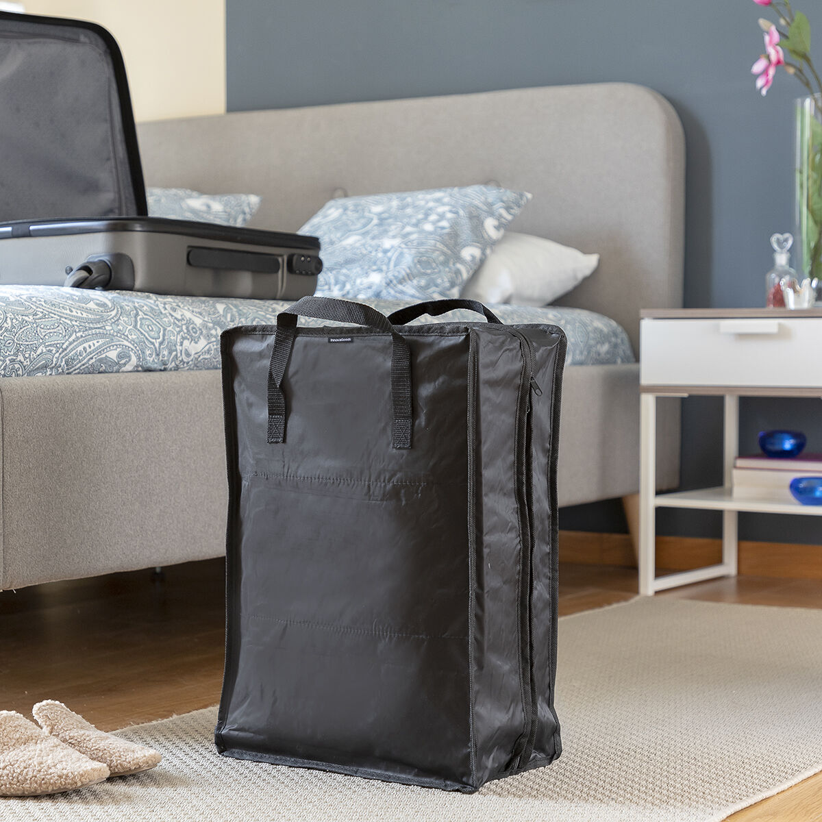 Eine schwarze Reisetasche steht in einem Zimmer mit Bett, ein Koffer liegt auf dem Bett. Nachttisch rechts.