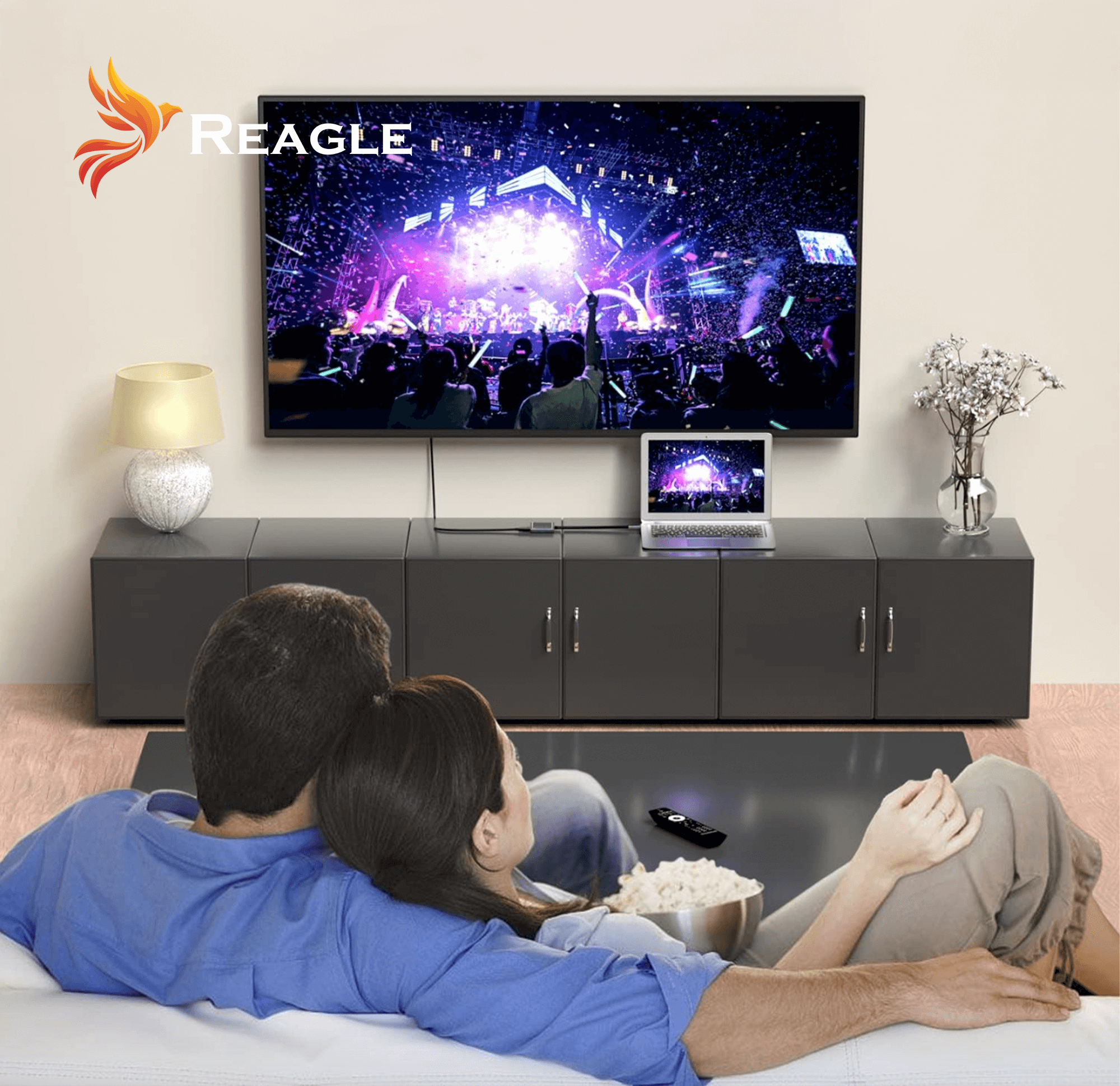 Coppia che guarda un film su una grande TV. Ciotola di popcorn sul tavolino. Logo Reagle.