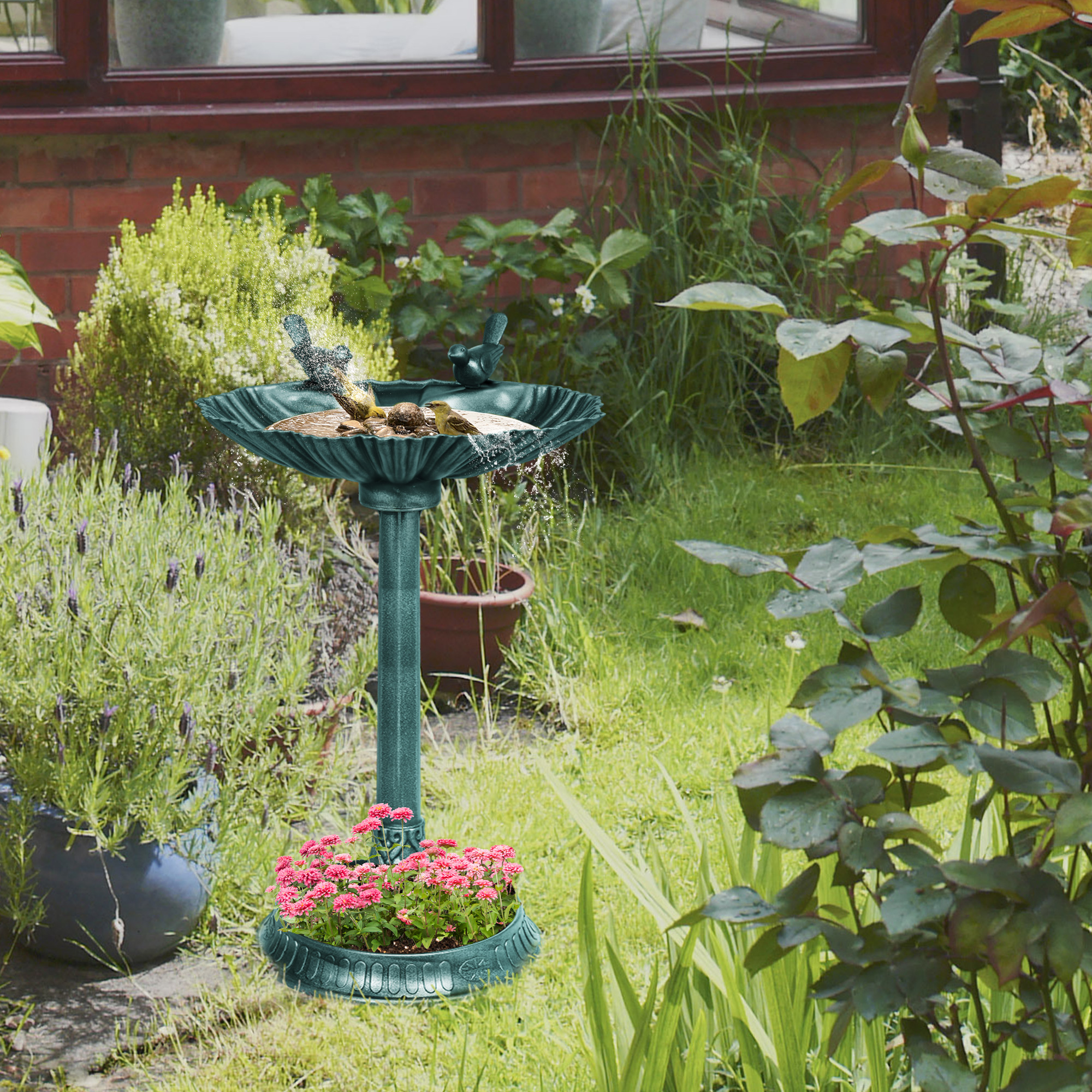 Vogelbad im Garten mit Grün, Blumen und Backsteinmauer im Hintergrund.