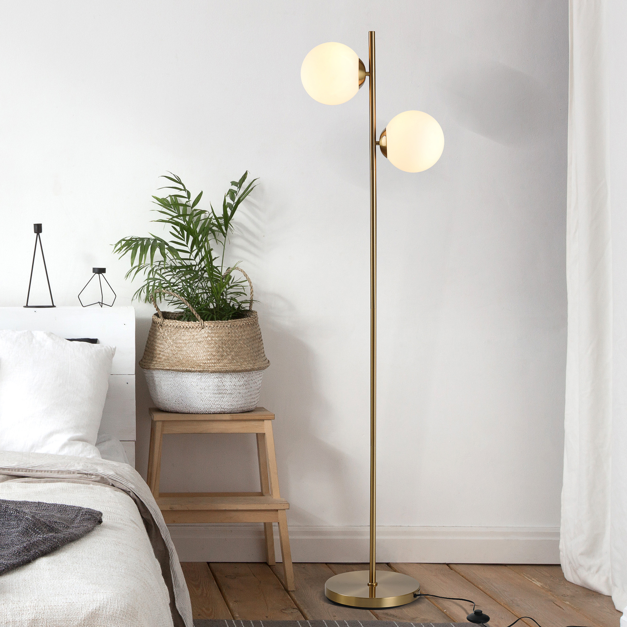 Eine goldene Stehlampe mit zwei Kugelleuchten. Ein Bett und eine Pflanze im Raum.