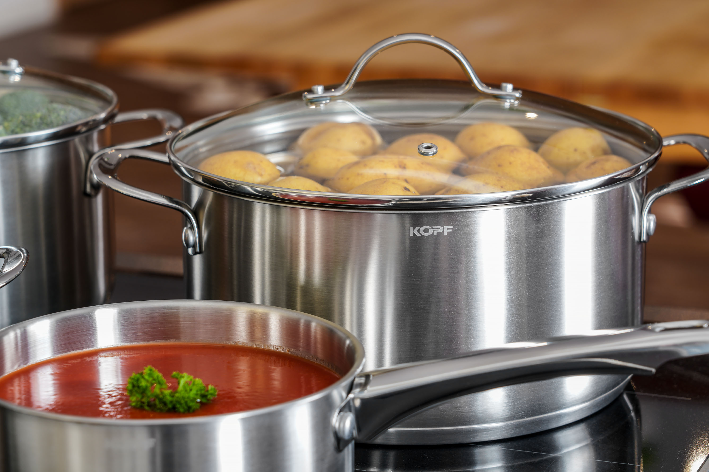 Edelstahltöpfe auf einem Kochfeld, einer mit Kartoffeln, der andere mit Tomatensauce. Ein Glasdeckel liegt auf dem Topf.