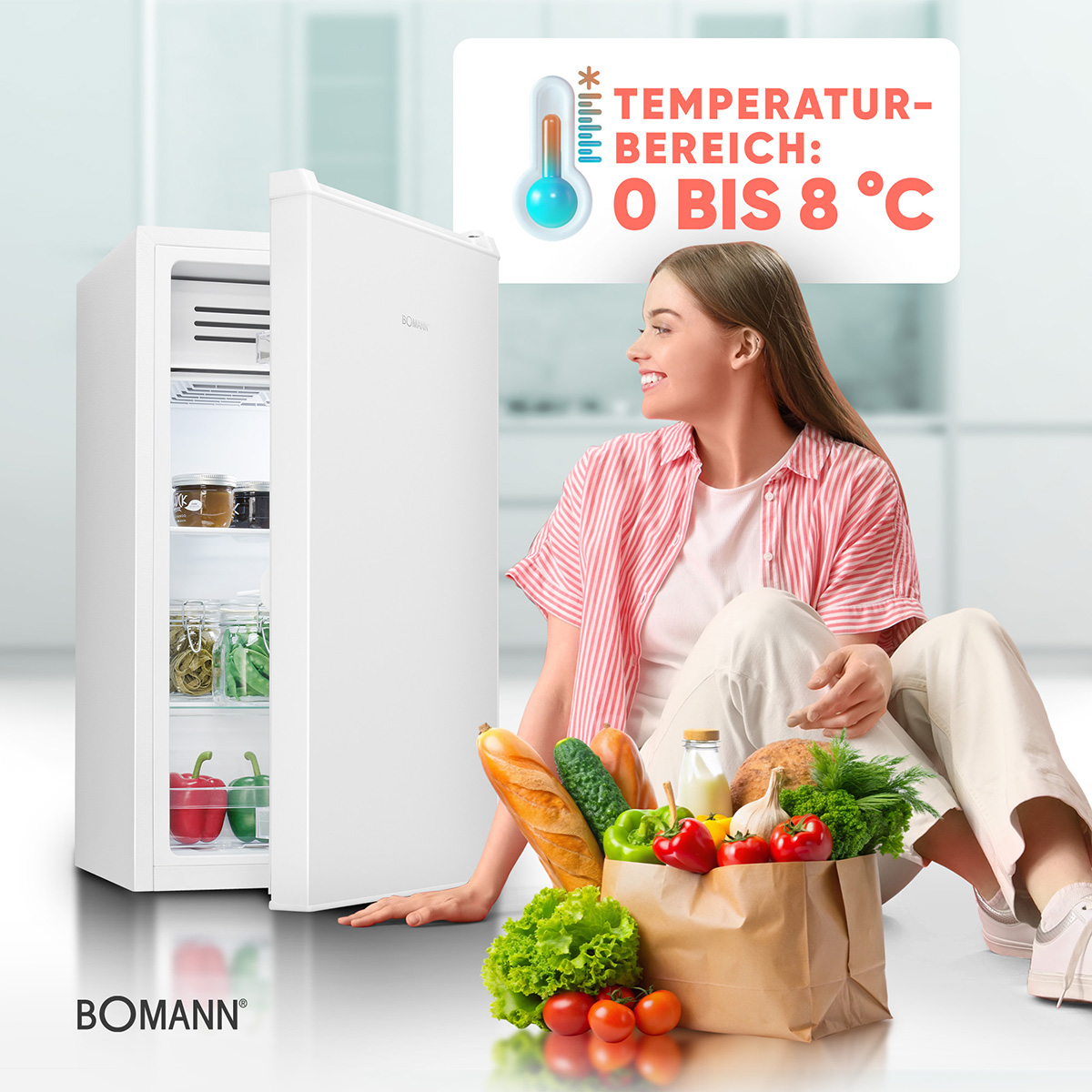 Frau mit Lebensmitteln sitzt neben einem weißen Kühlschrank.
