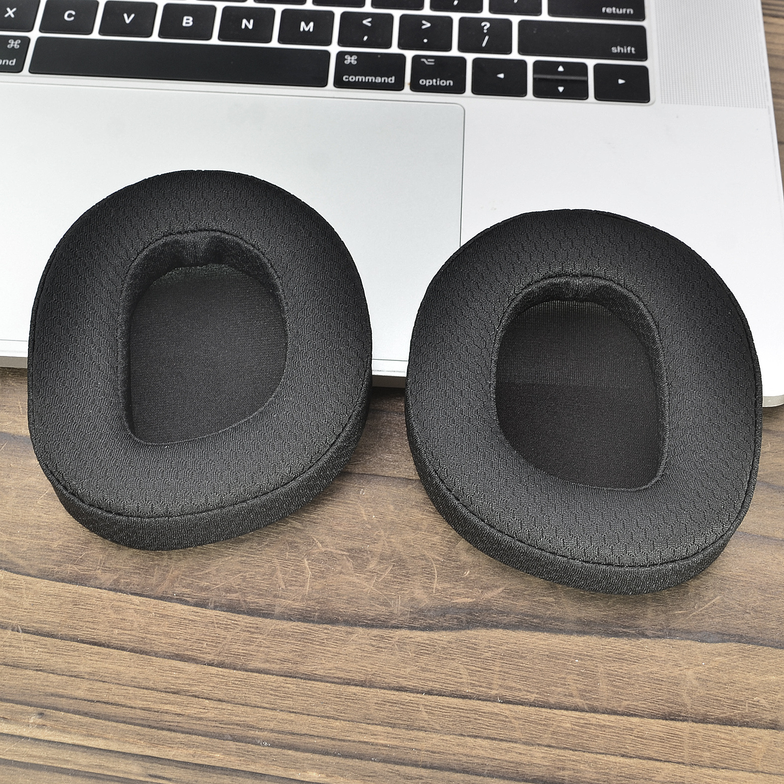 Due cuscinetti auricolari neri per cuffie su una scrivania di legno. Un laptop sullo sfondo.