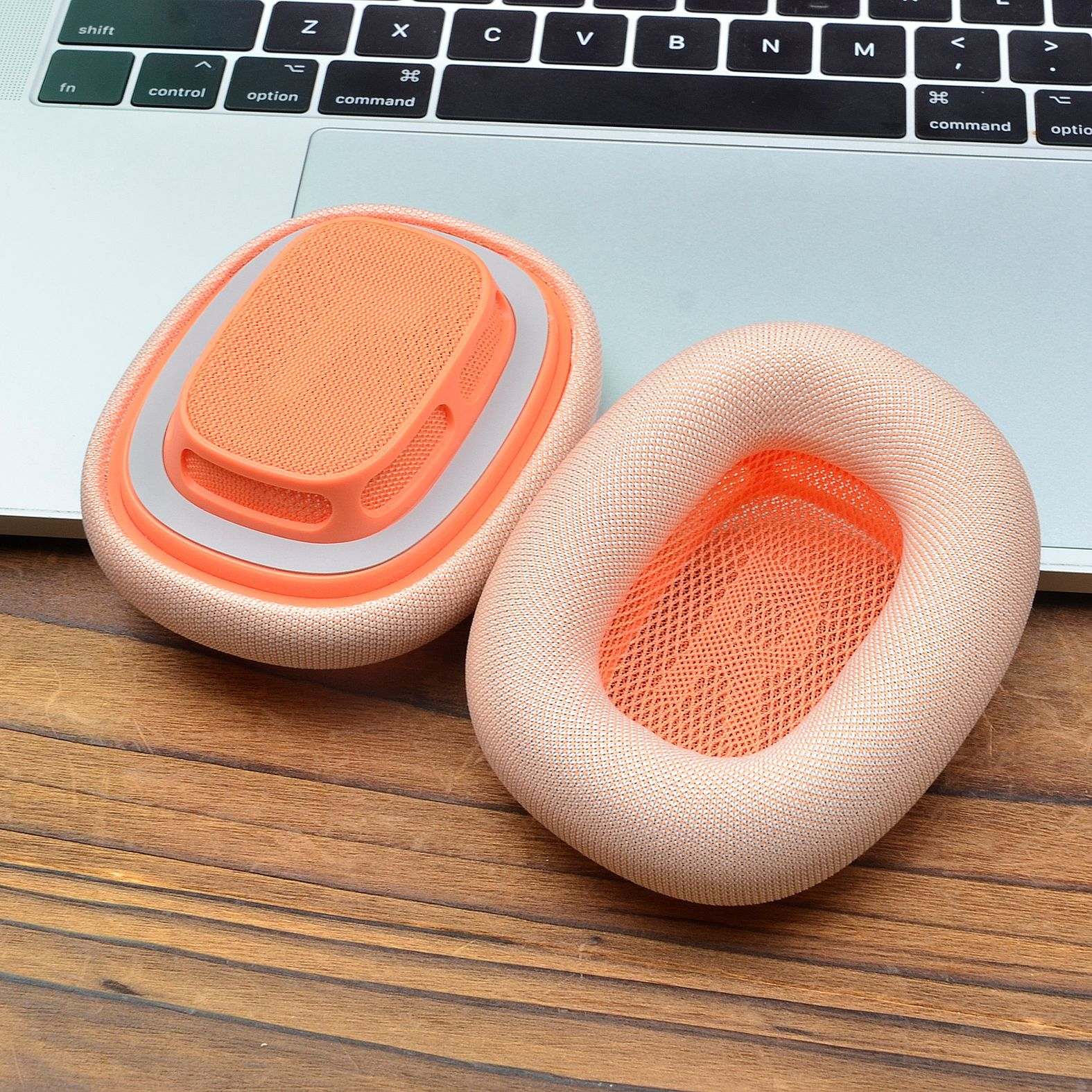 Zwei pfirsichfarbene Kopfhörer-Ohrpolster mit orangefarbenem Mesh-Innenraum, Laptop und Holz.