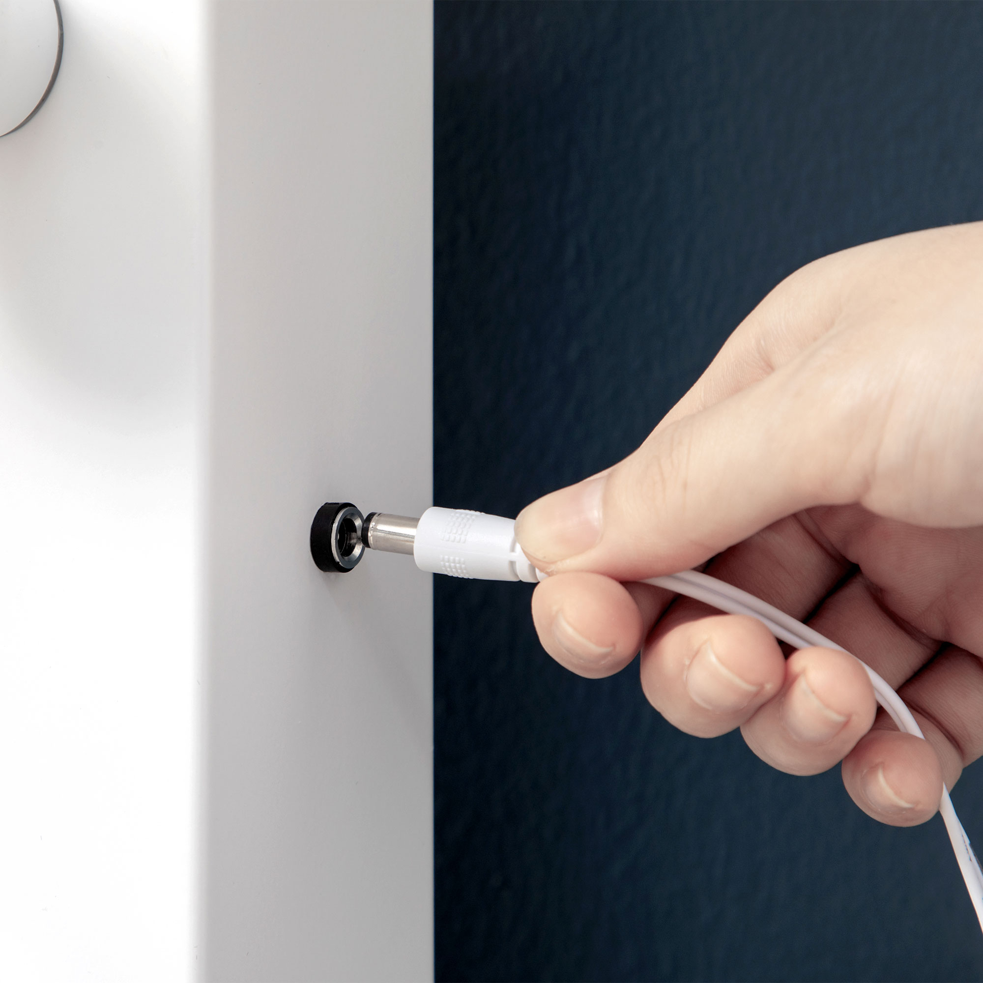 Hand, die ein weißes Ladekabel in einen kleinen schwarzen Ladeanschluss auf weißer Oberfläche steckt.