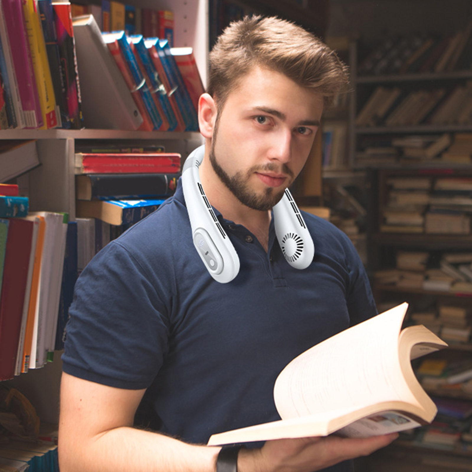 Mann in Bibliothek mit Nackenventilator, hält Buch und schaut in die Kamera. Bücherregale füllen den Hintergrund.