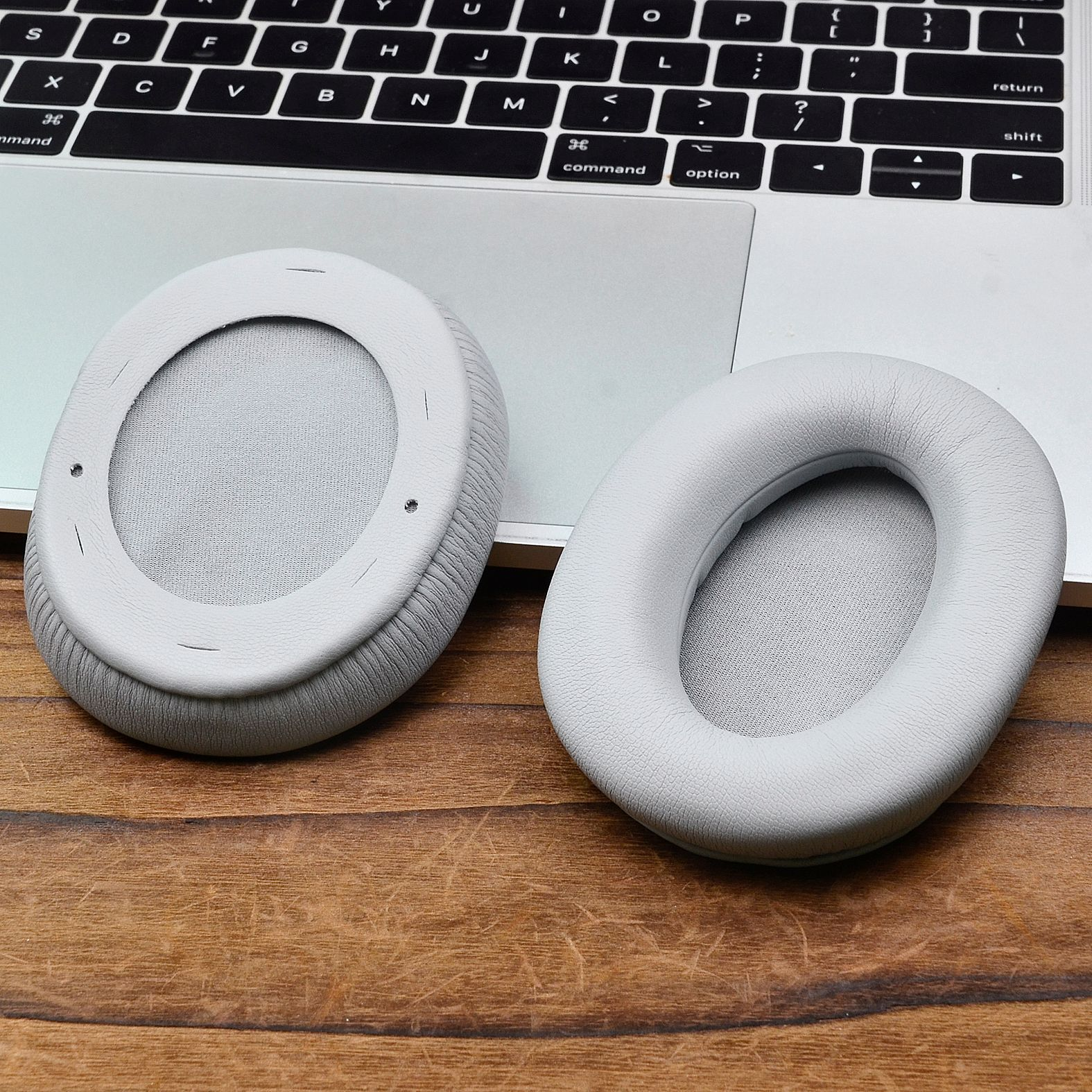Due cuscinetti auricolari grigio chiaro per cuffie su un tavolo di legno. Un laptop è visibile sullo sfondo.
