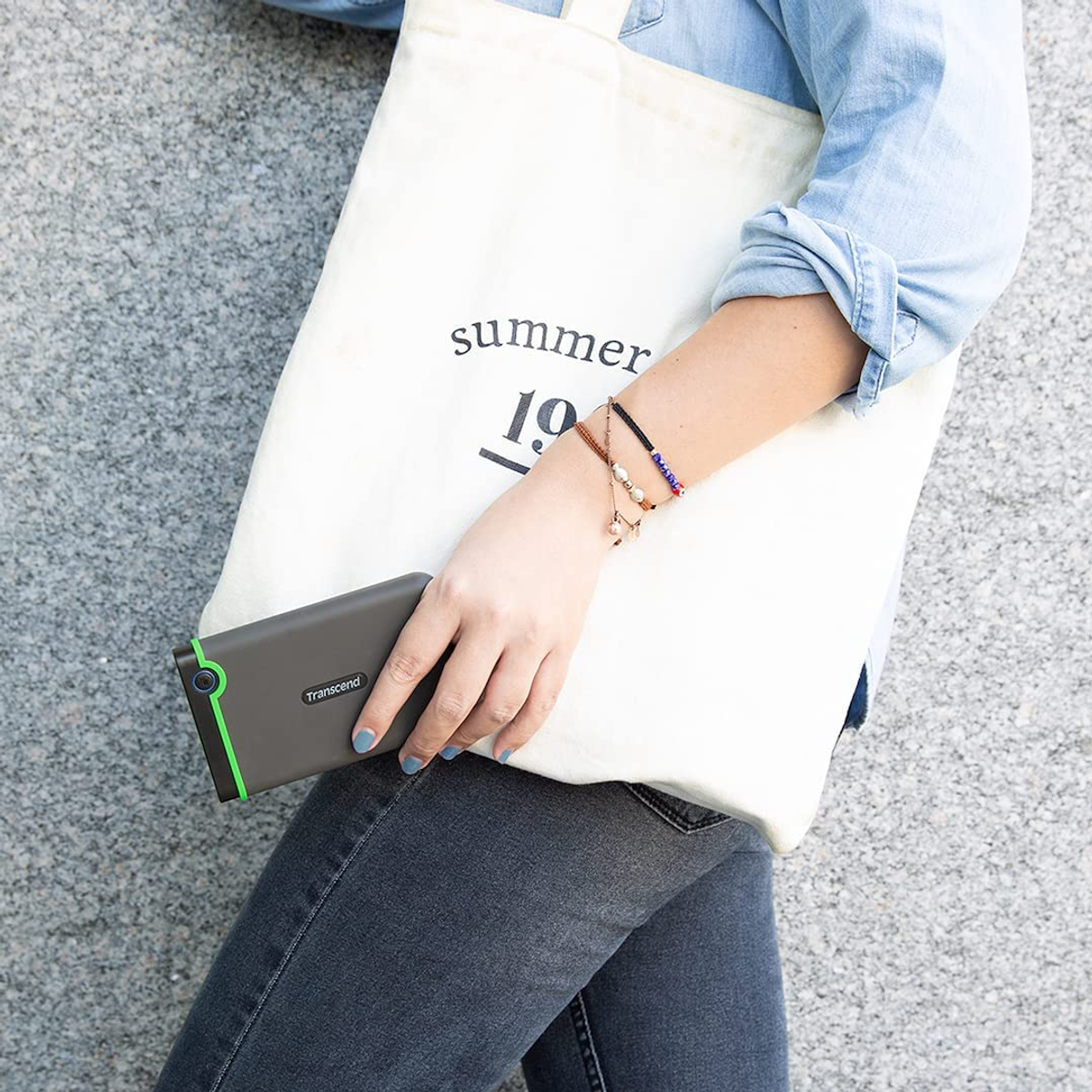 Kobieta trzyma torbę i zewnętrzny dysk twardy. Torba ma tekst, napęd jest szary.