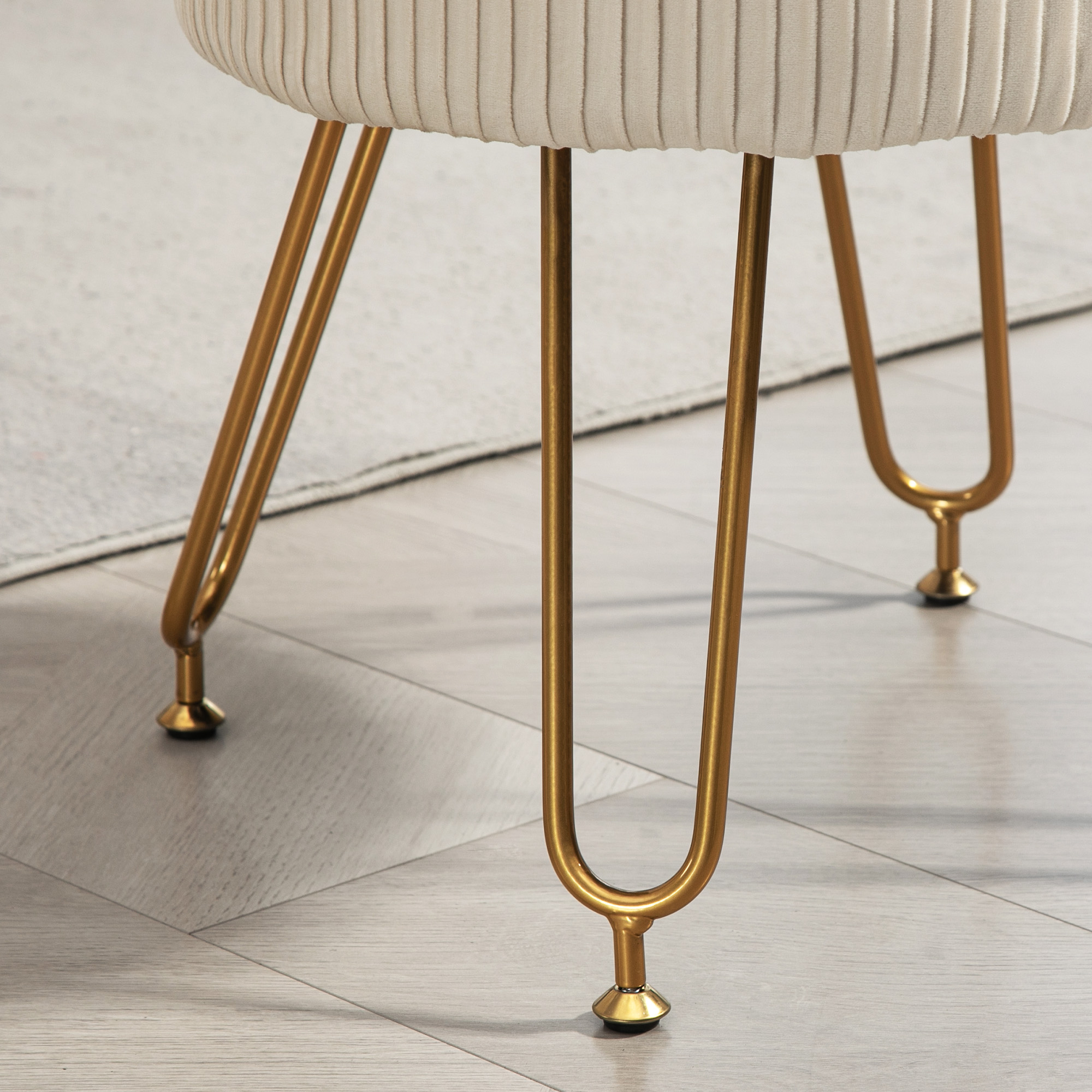 Ein beiger, gerippter Stoffhocker mit goldfarbenen, gebogenen Metallbeinen auf hellem Boden.