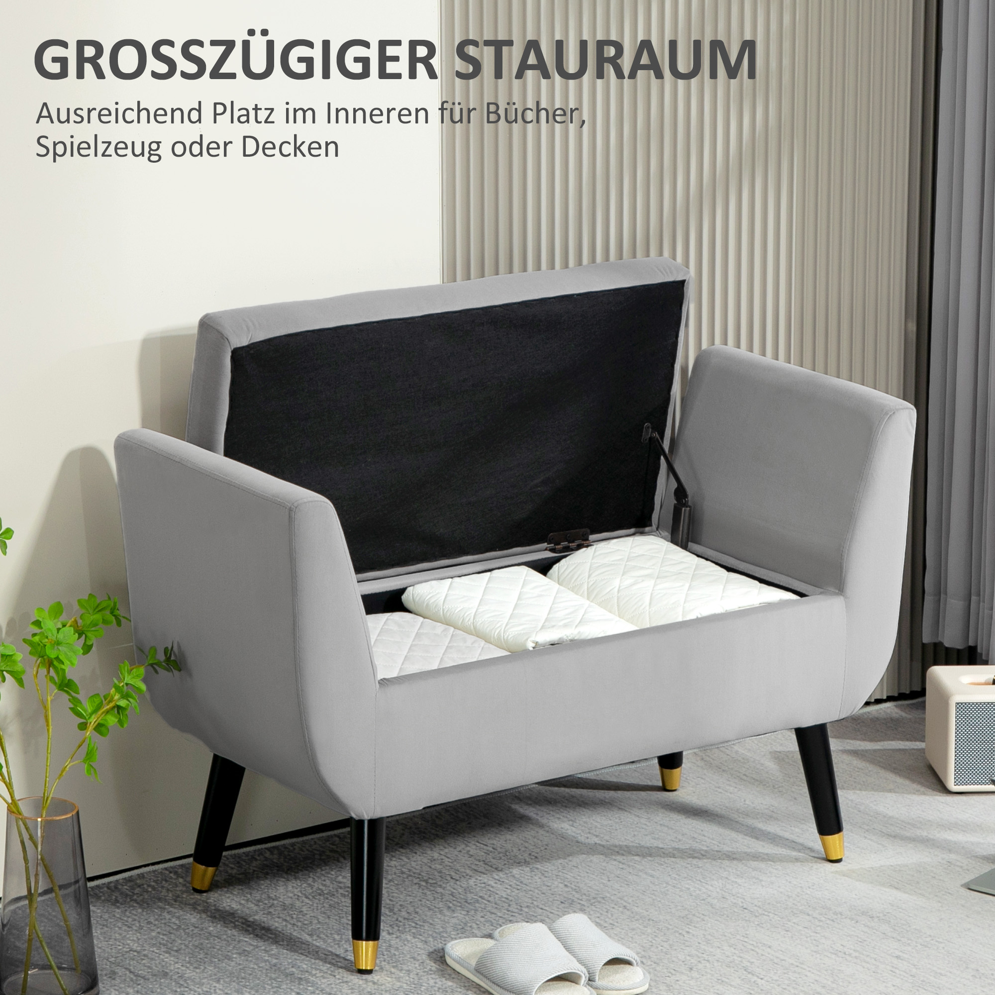 Graue Aufbewahrungsbank mit offenem Deckel, zeigt weiße Kissen, mit schwarzen Beinen und goldenen Akzenten.