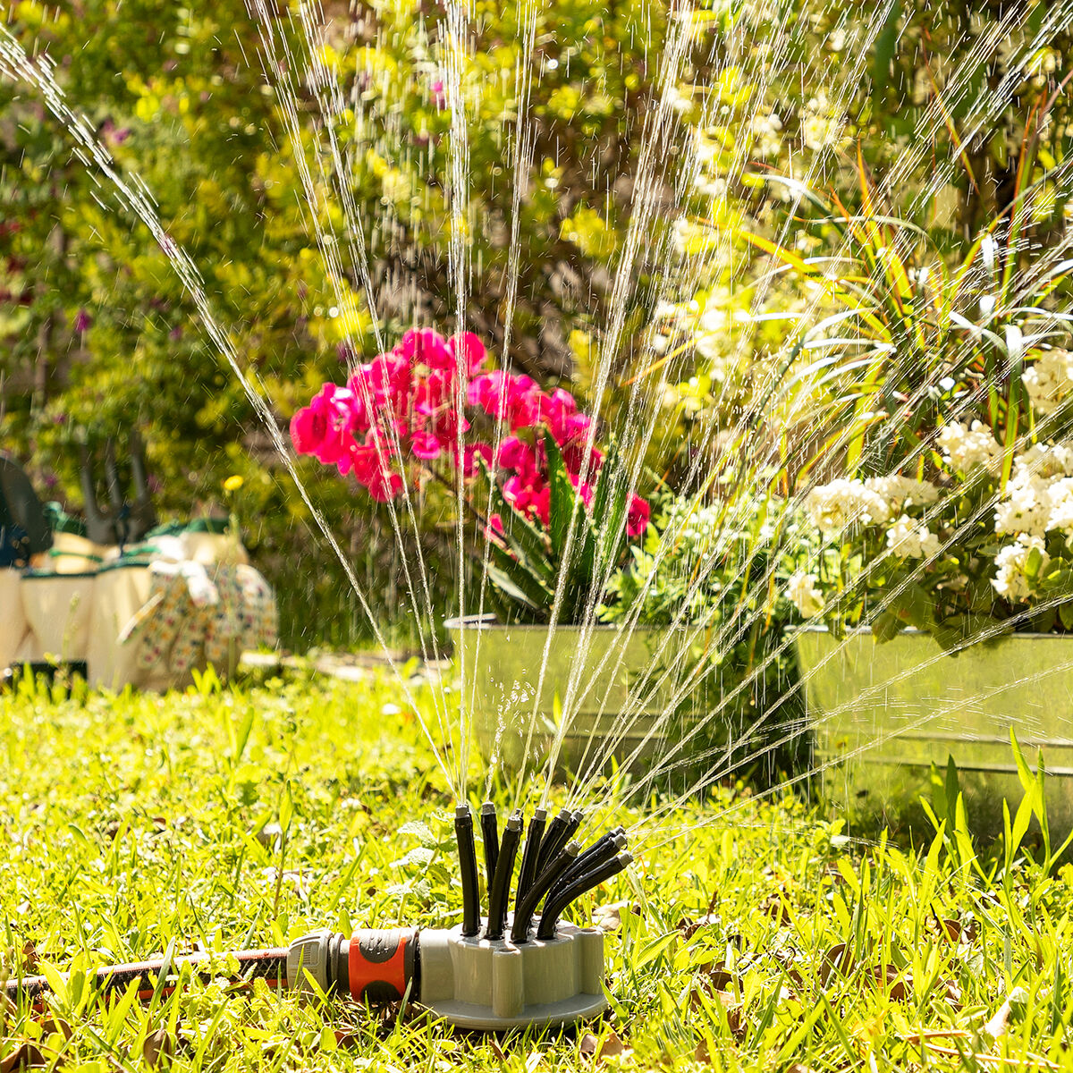 Rasensprenger bewässert grünen Rasen. Rosa Blumen und grüne Pflanzen im Hintergrund.