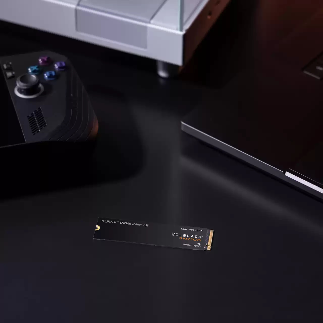 SSD NVMe negra con texto blanco y dorado, cerca de consola, portátil y mando, sobre una superficie negra.