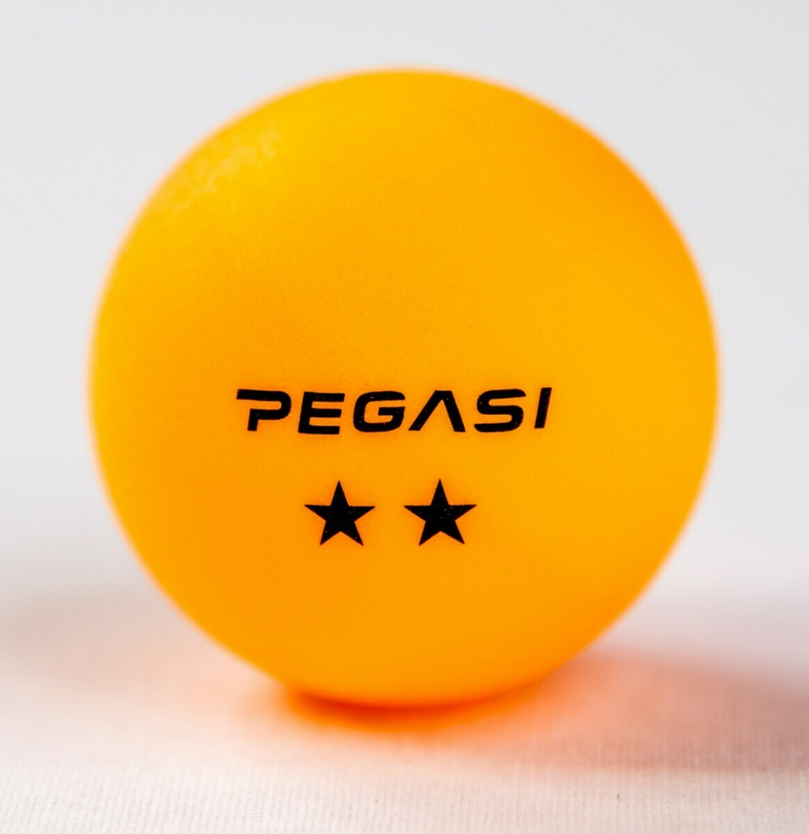 An orange ping pong ball with 'PEGASI' and two stars printed on it, over a white backdrop.