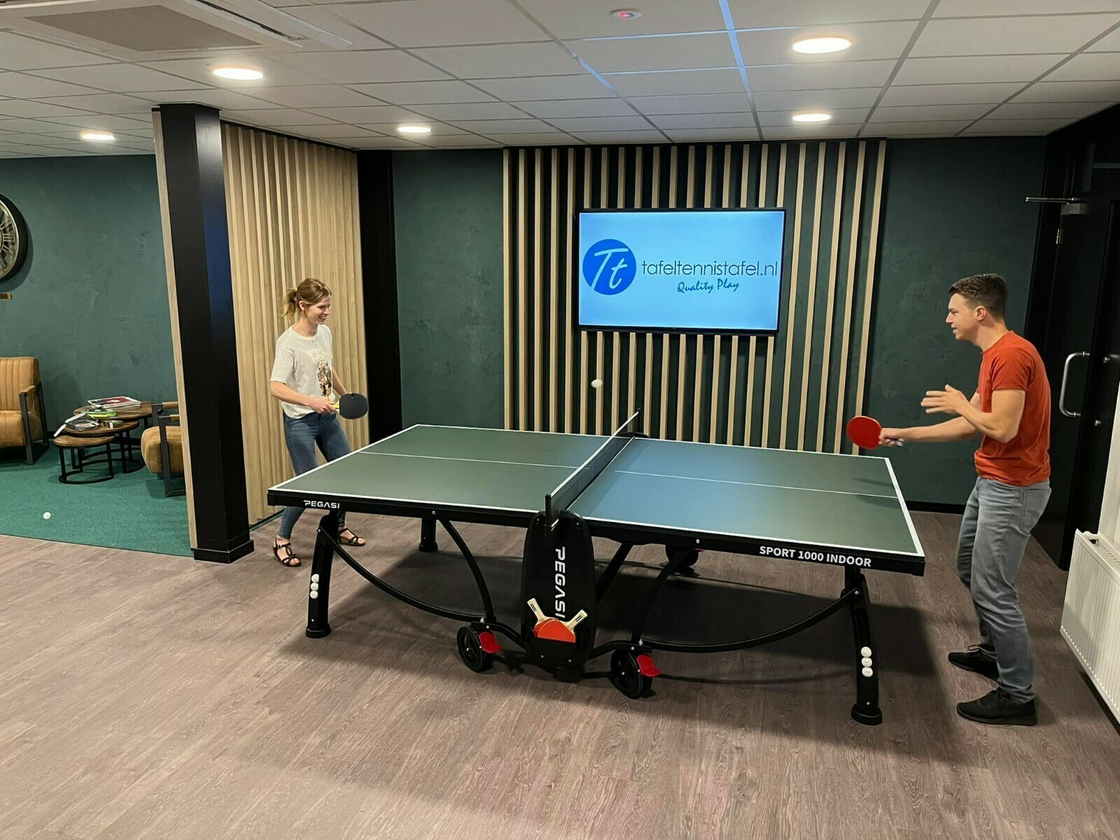 Two people playing table tennis. A ball is in the air.