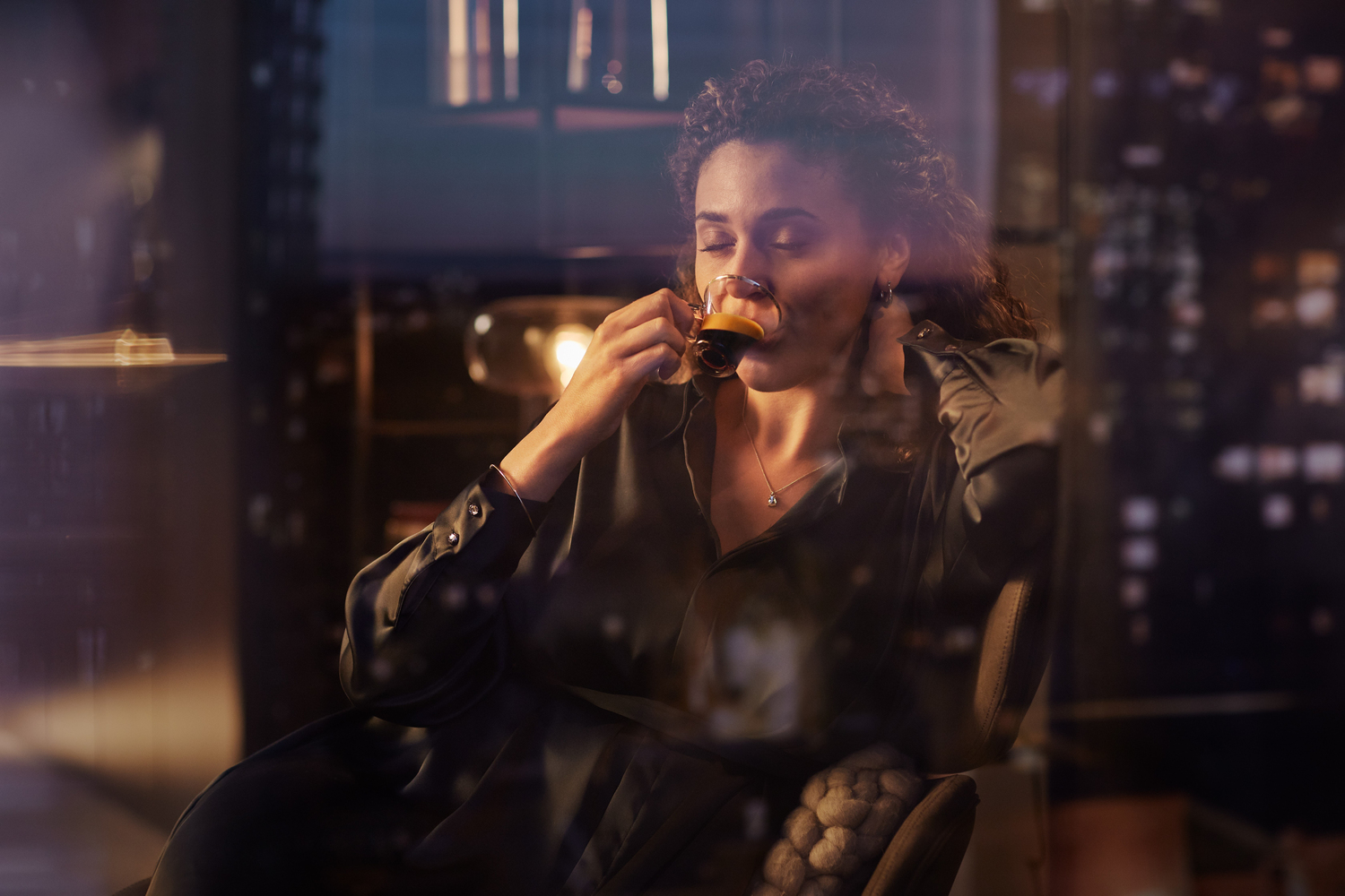 Woman drinking coffee near a window at night. City lights in background.