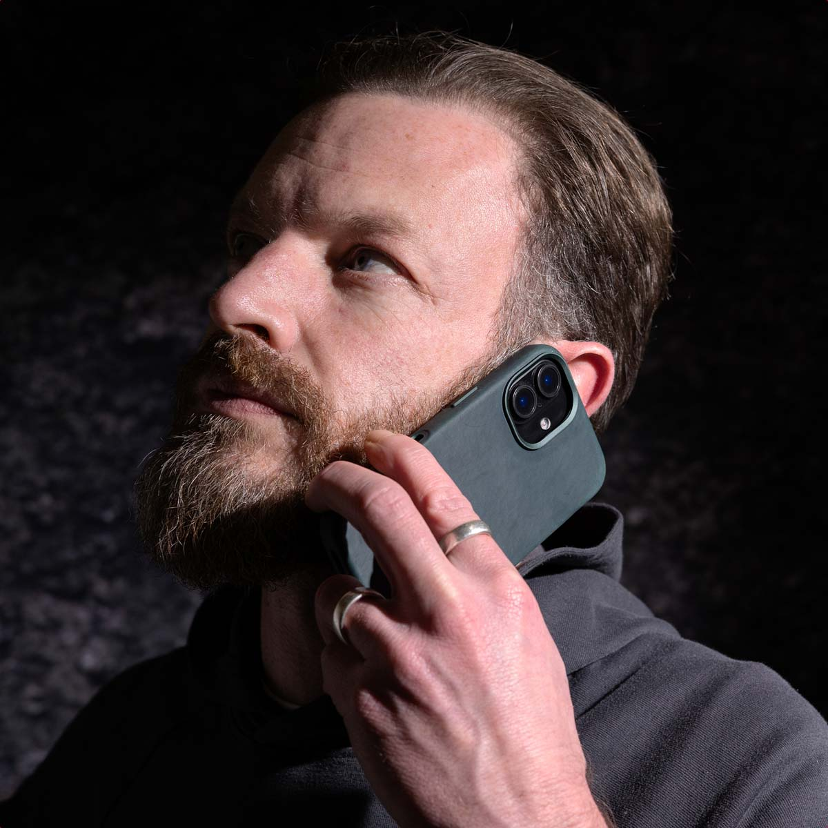 Man met baard gebruikt smartphone, kijkt omhoog, donkere achtergrond.