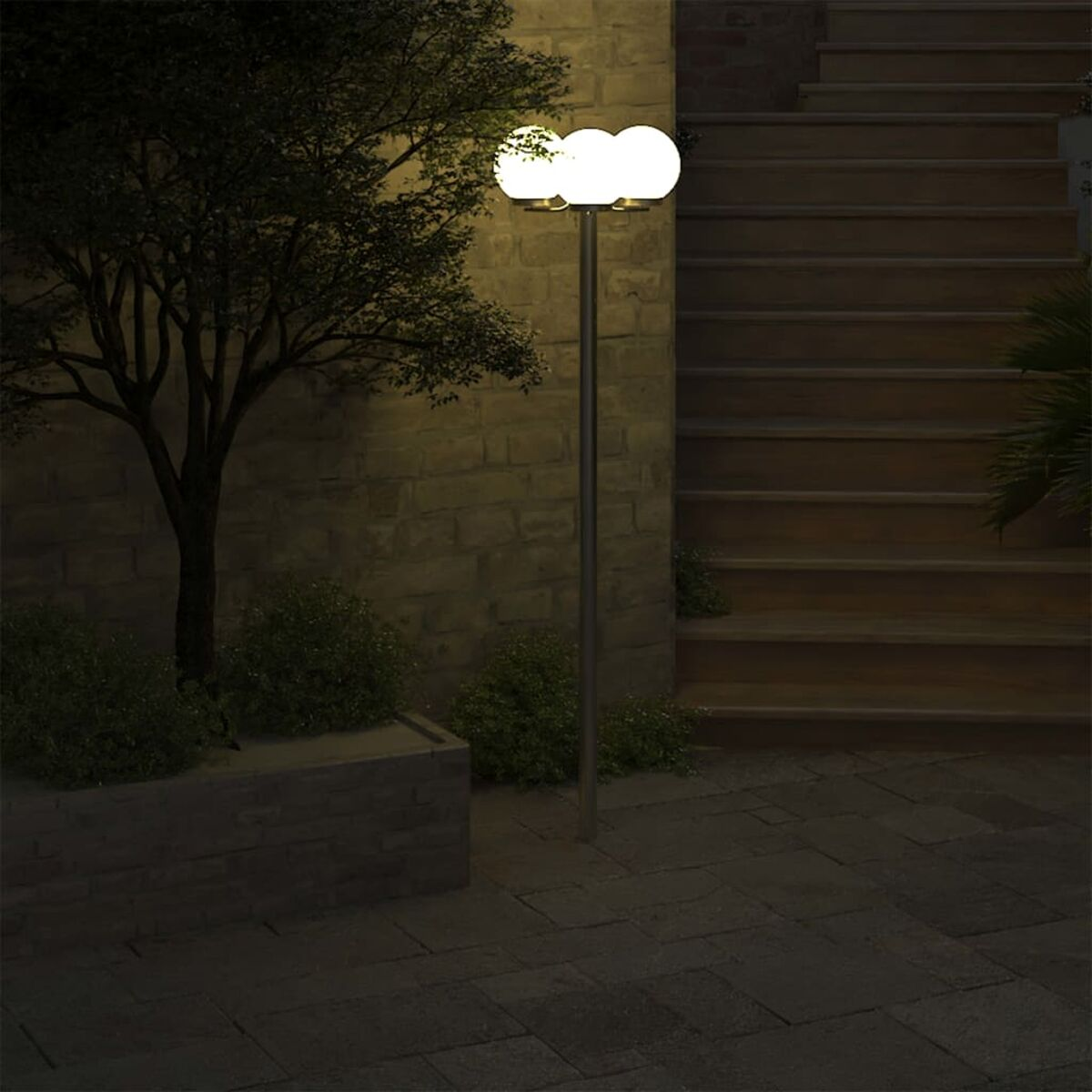 Un lampione esterno illuminato con tre globi bianchi. Scale e un muro di mattoni sullo sfondo.