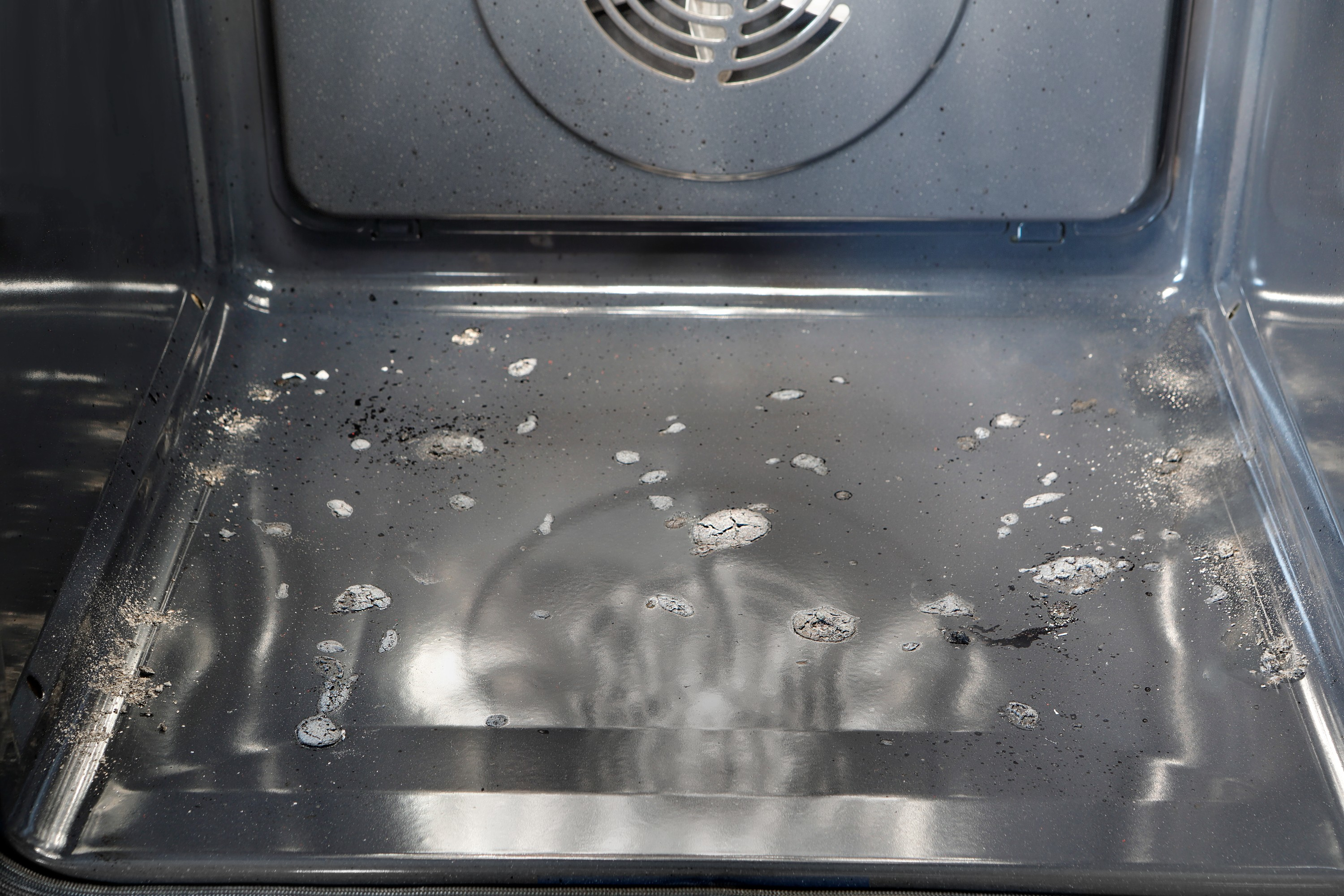 Dentro un forno sporco. Il fondo è grigio con residui di cibo, macchie. È visibile una ventola.
