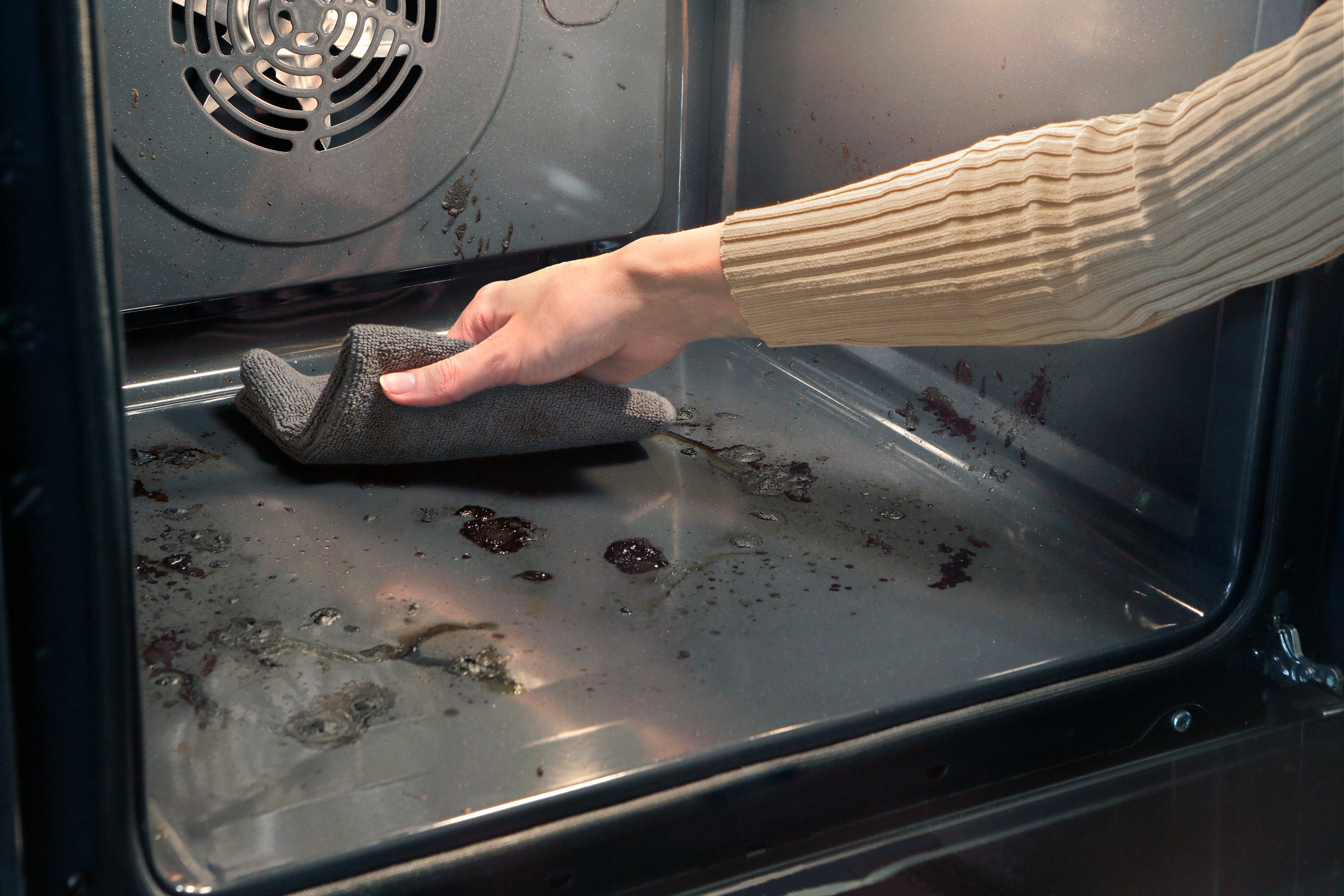 Mano che pulisce l'interno sporco del forno con un panno grigio, mostrando residui di cibo.