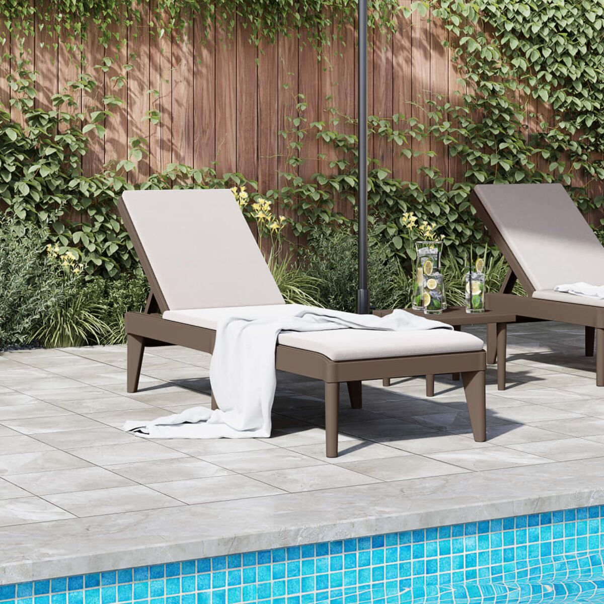 Zwei braune Sonnenliegen auf einer gefliesten Terrasse am Pool. Es gibt Getränke und ein Handtuch.