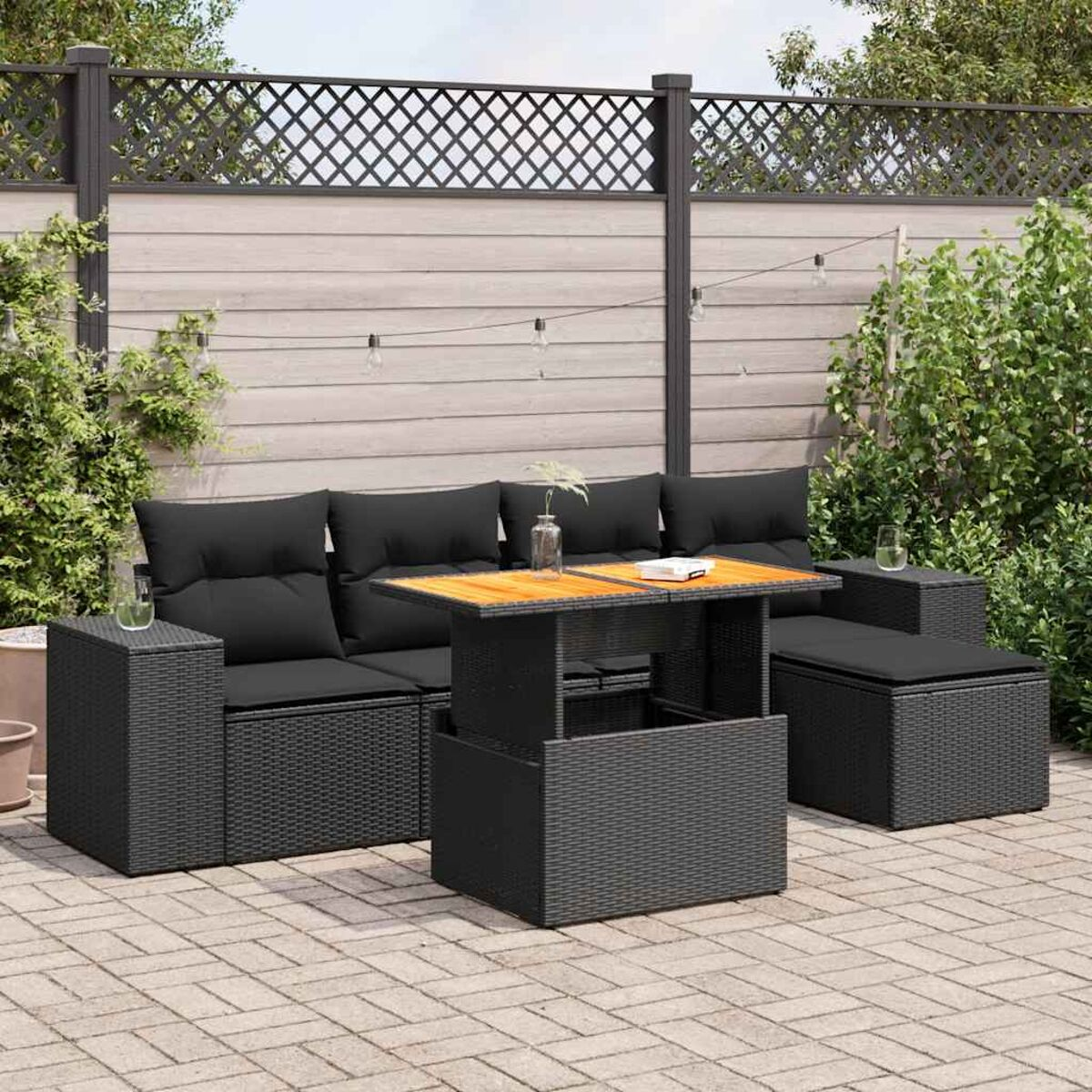 Schwarzes Gartenmöbel-Set mit schwarzen Kissen und einem Holztisch. Ziegelterrasse, Zaun und Pflanzen im Hintergrund.