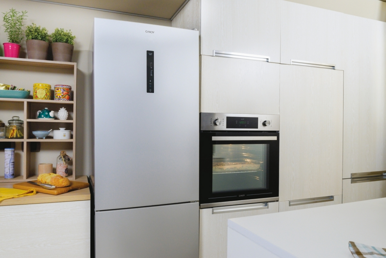 Una scena di cucina con frigorifero, forno e armadi. È visibile una mensola con piante e pane.