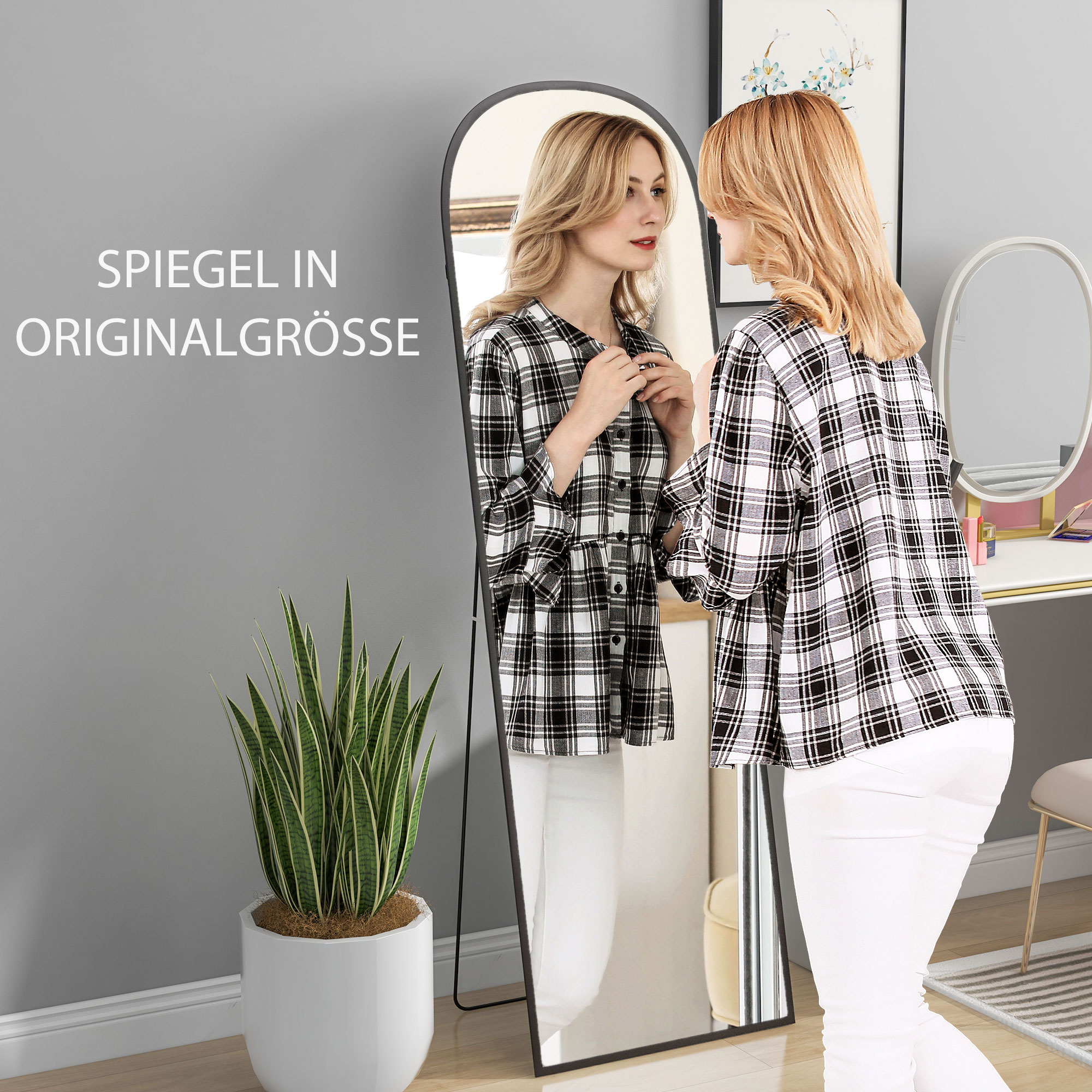Frau betrachtet sich in einem großen Spiegel. Sie trägt ein kariertes Hemd und weiße Hosen.