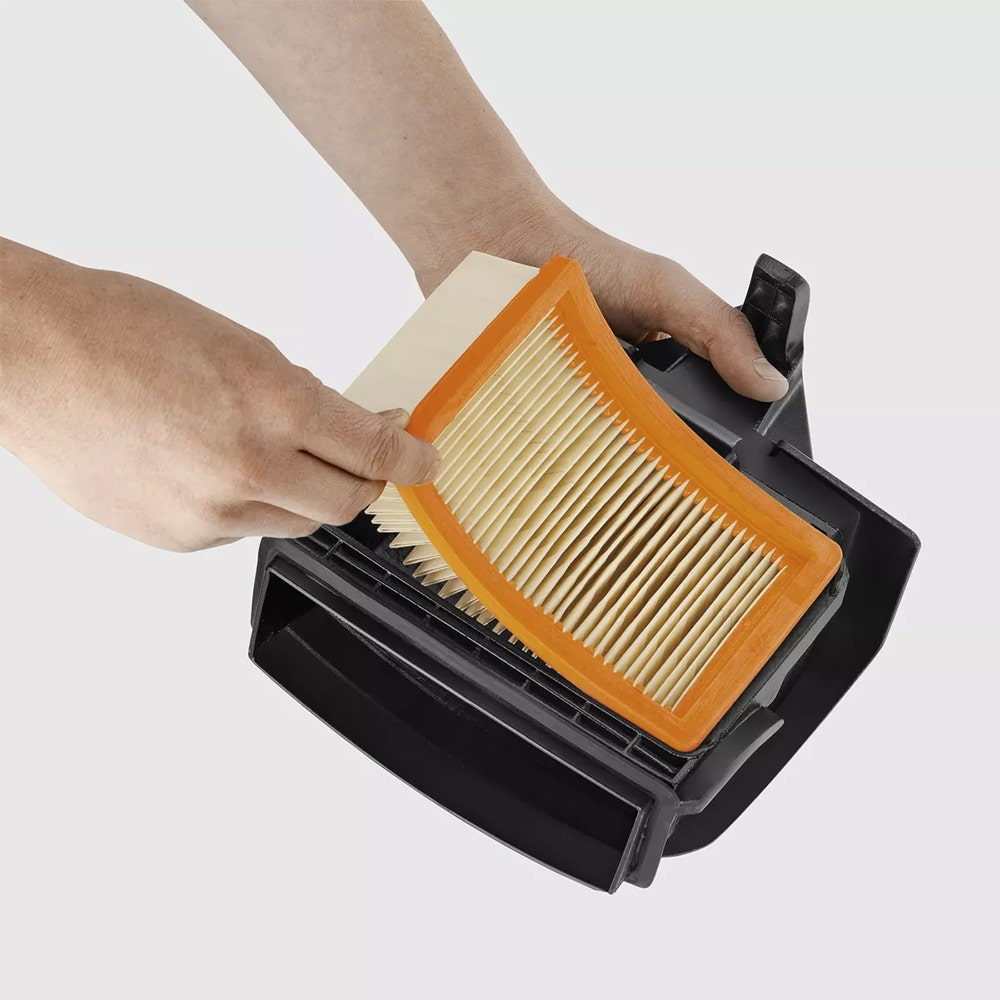 Mani che rimuovono un filtro dell'aria da un alloggiamento nero. Il filtro è arancione e beige. Sfondo bianco.