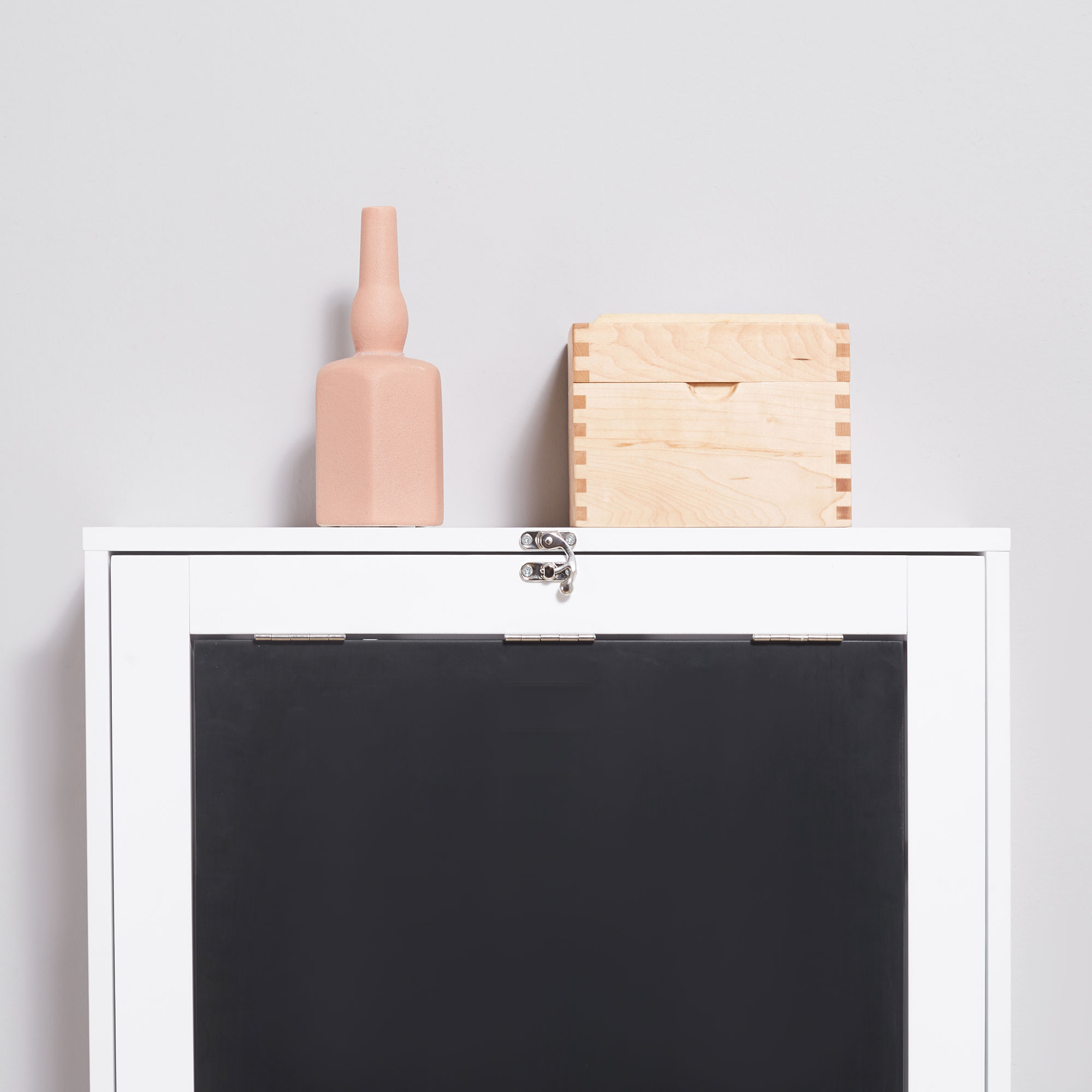 Ein weißer Schrank mit einer schwarzen Tafelklappe und einer rosa Vase und einer Holzkiste oben drauf.