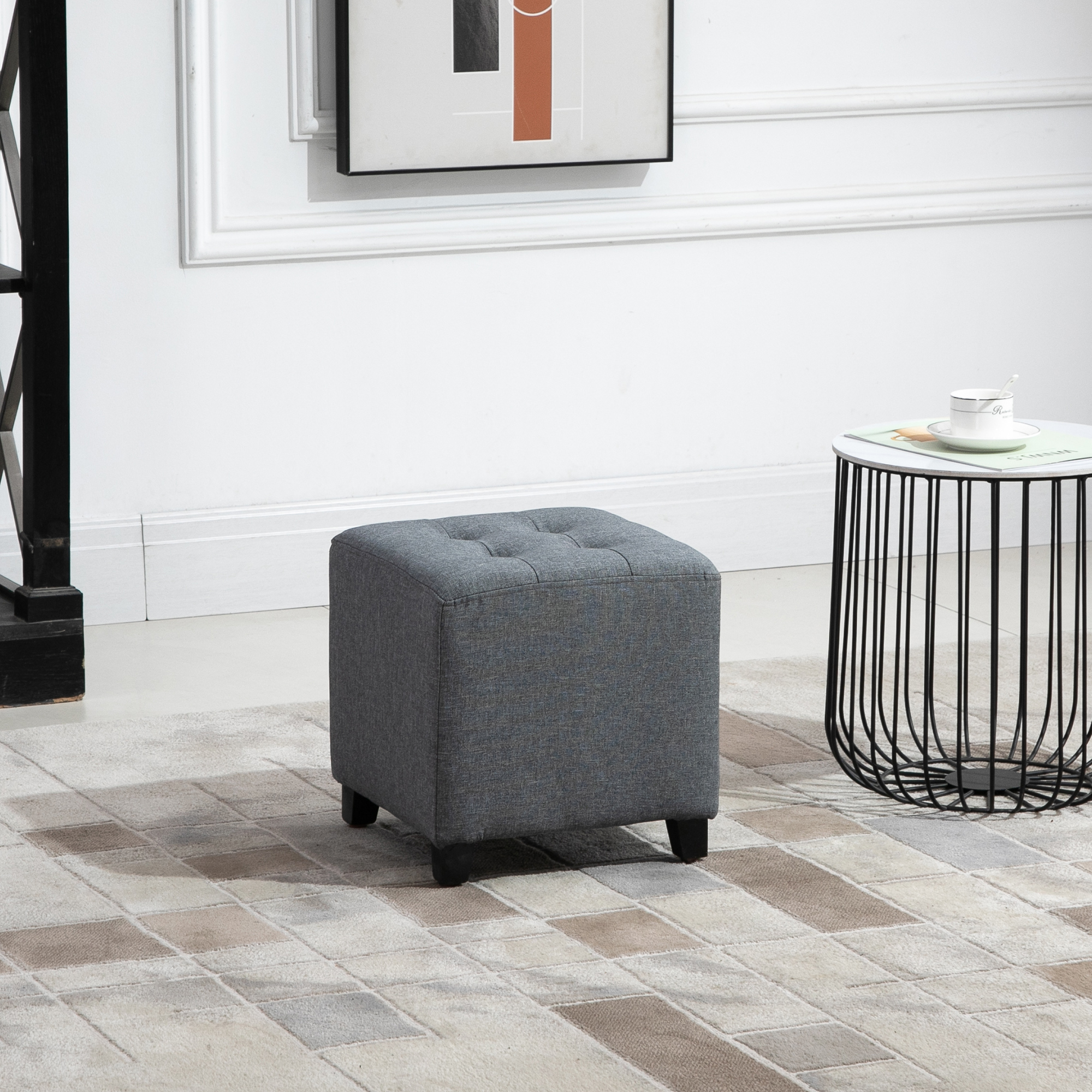 Ein grauer quadratischer Ottoman mit Knopfheftung auf einem gemusterten Teppich. Daneben steht ein schwarz-weißer Tisch.