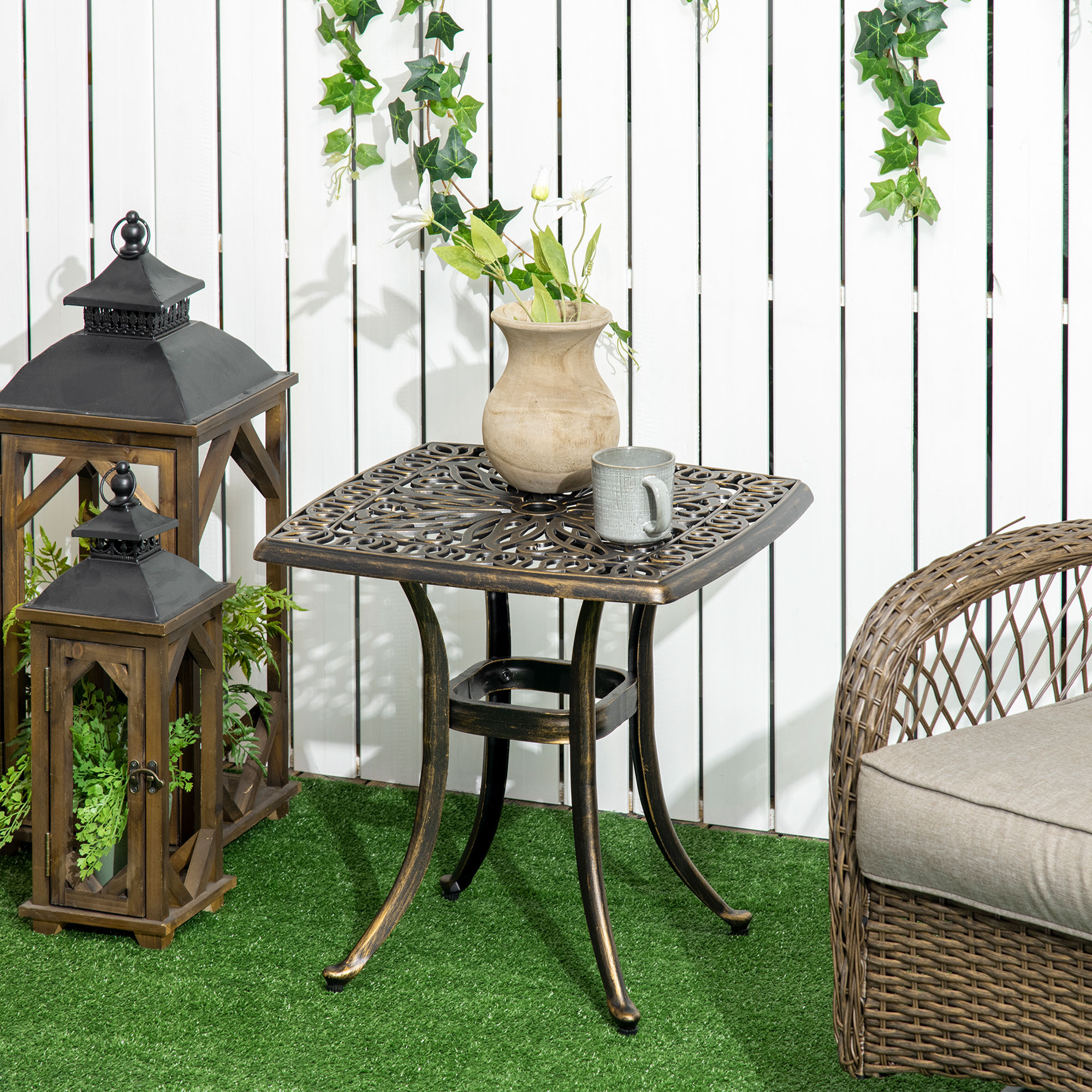 Quadratischer Metall-Gartentisch mit floralem Design. Zwei Laternen und eine Tasse auf grünem Gras.