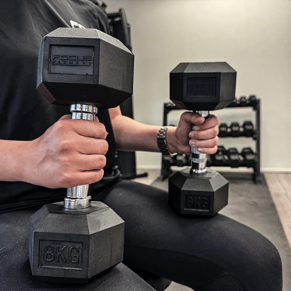 Person holding two black 8KG dumbbells. Focus Fitness logo is visible. Wearing black clothes and a watch.