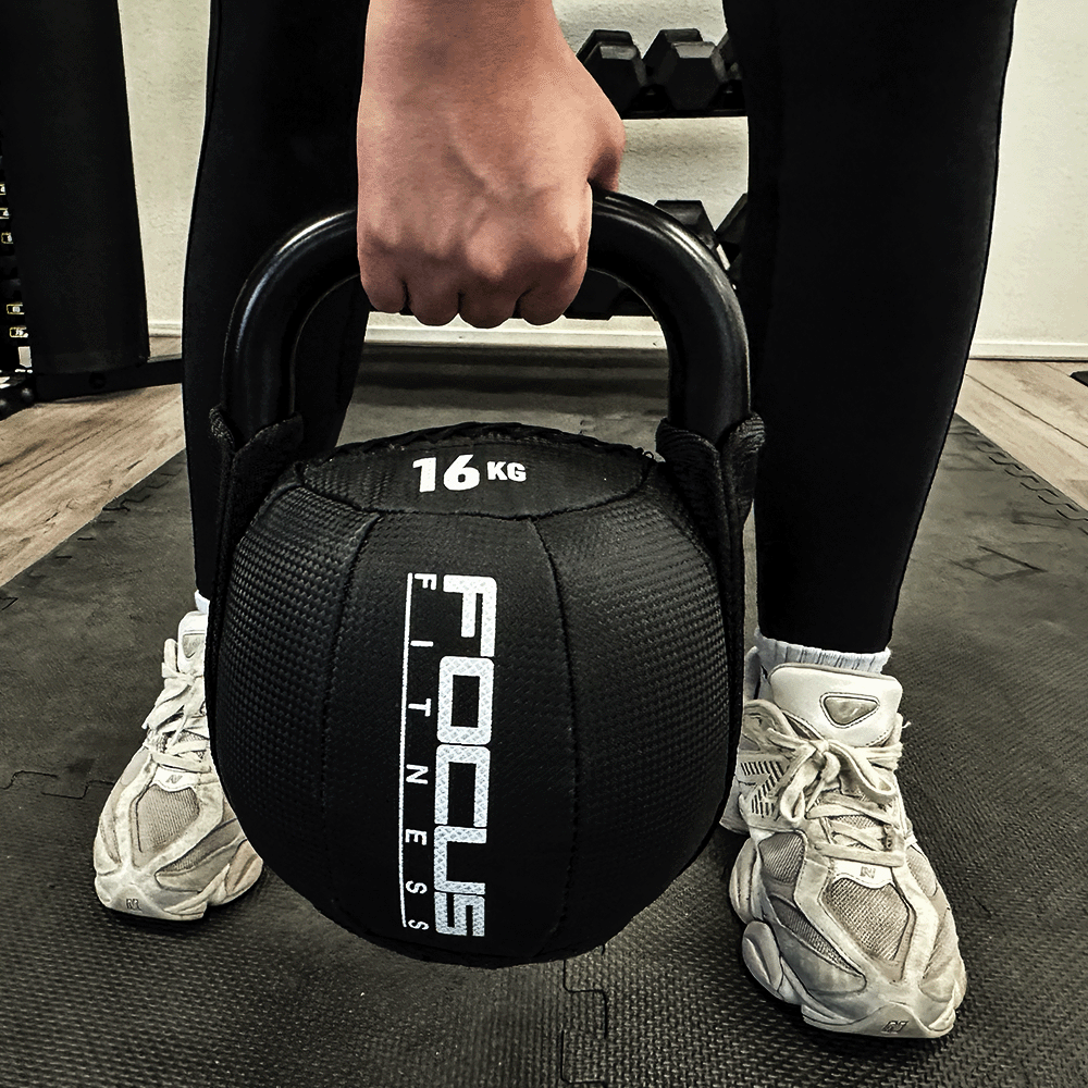 Person holding a black 16KG kettlebell. Focus Fitness logo is visible. Wearing black leggings and white shoes.