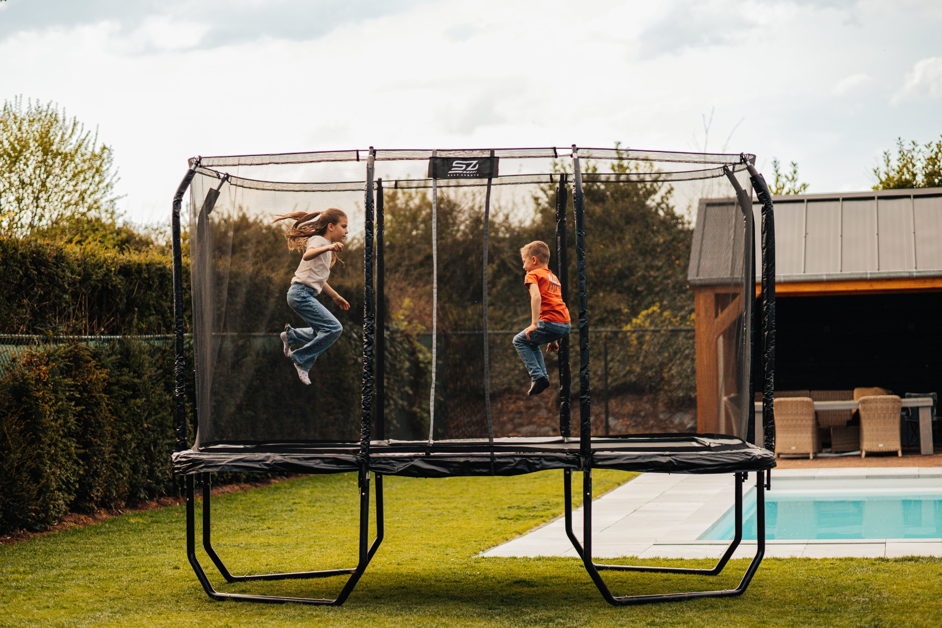 Twee kinderen springen op een grote trampoline in een achtertuin. Blauwe lucht. Groen gras en een zwembad.