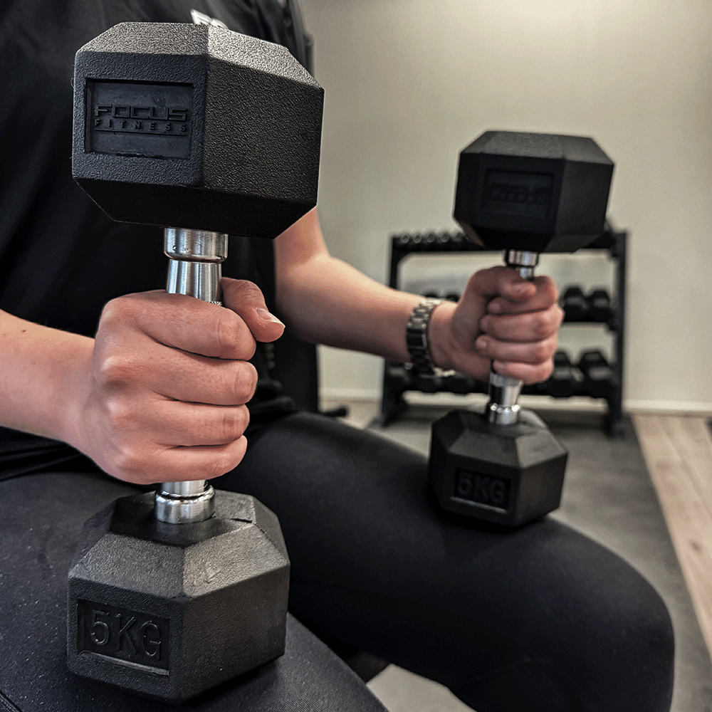 Person holding two black 5KG dumbbells. Focus Fitness logo visible. Wearing black clothes and a watch.