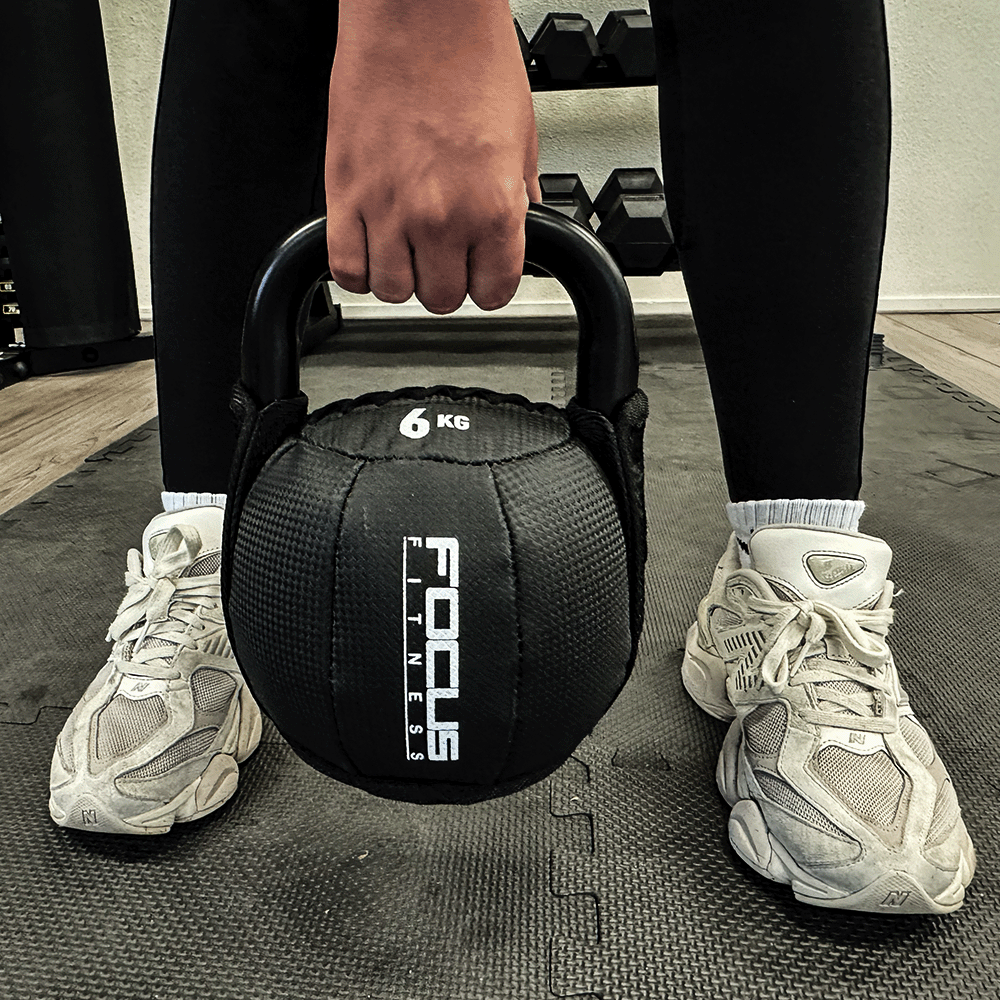 Person holding a black 6 KG kettlebell in a gym, wearing white sneakers.