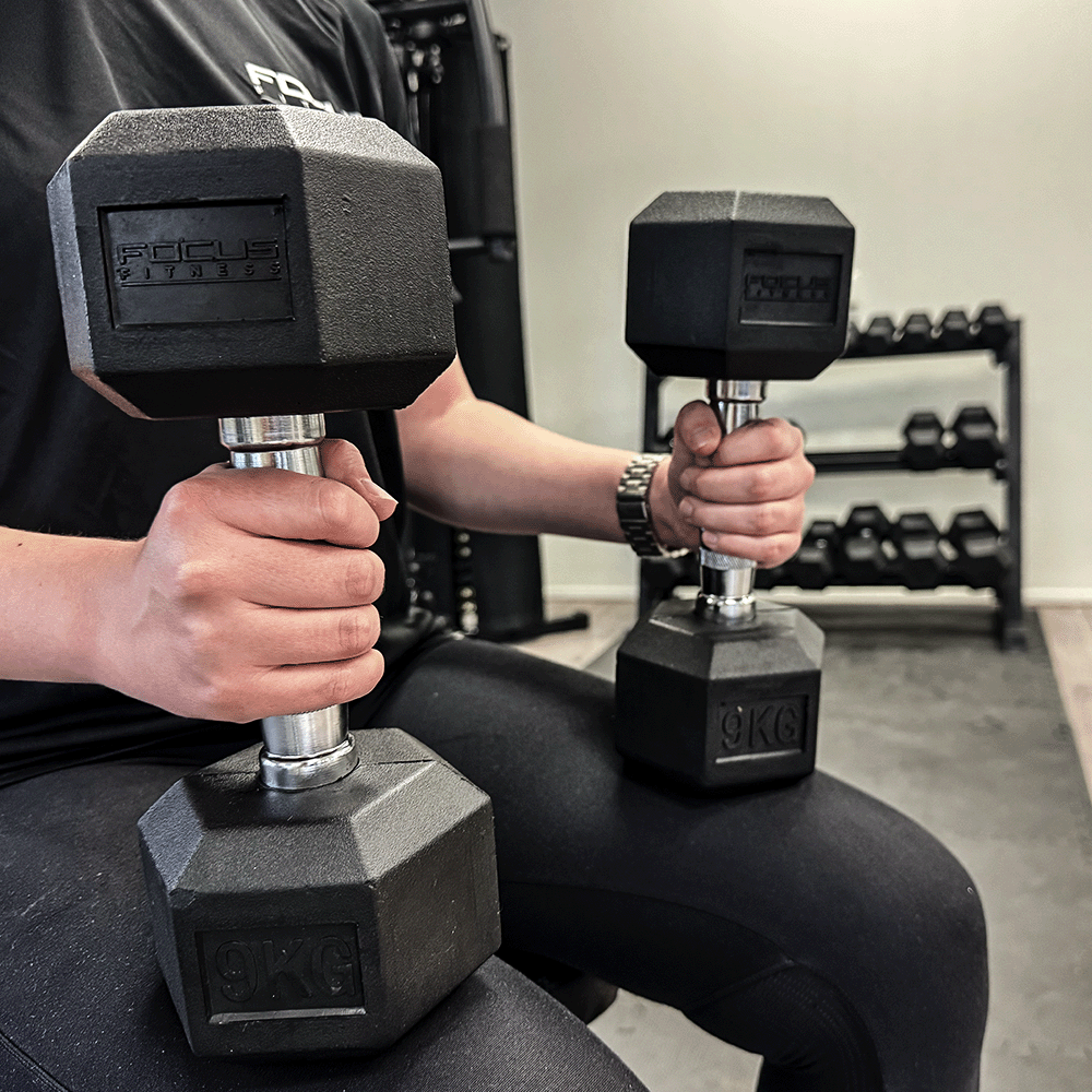Person holding two 9 KG black dumbbells in a gym setting.