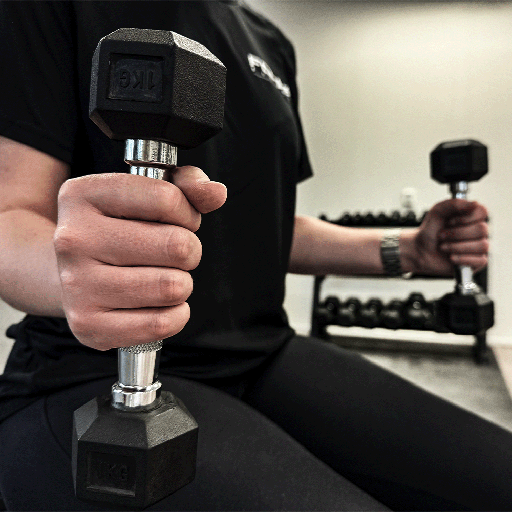 Person holding two black dumbbells, one 1 KG, in a gym.