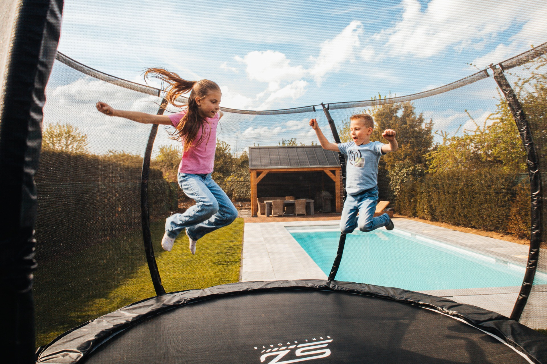 Twee kinderen springen vrolijk op een trampoline in een zonnige buitenomgeving. Blauwe lucht en groen gras.