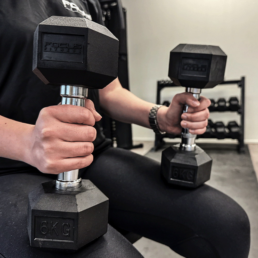 Person holding two 6 KG black dumbbells in a gym setting.